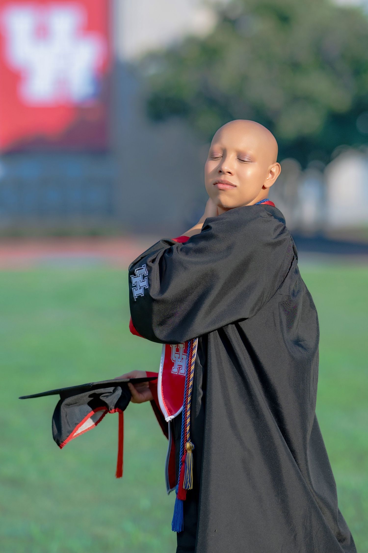 Bald graduate in black cap and gown posing confidently on a green field with UH logo in background.