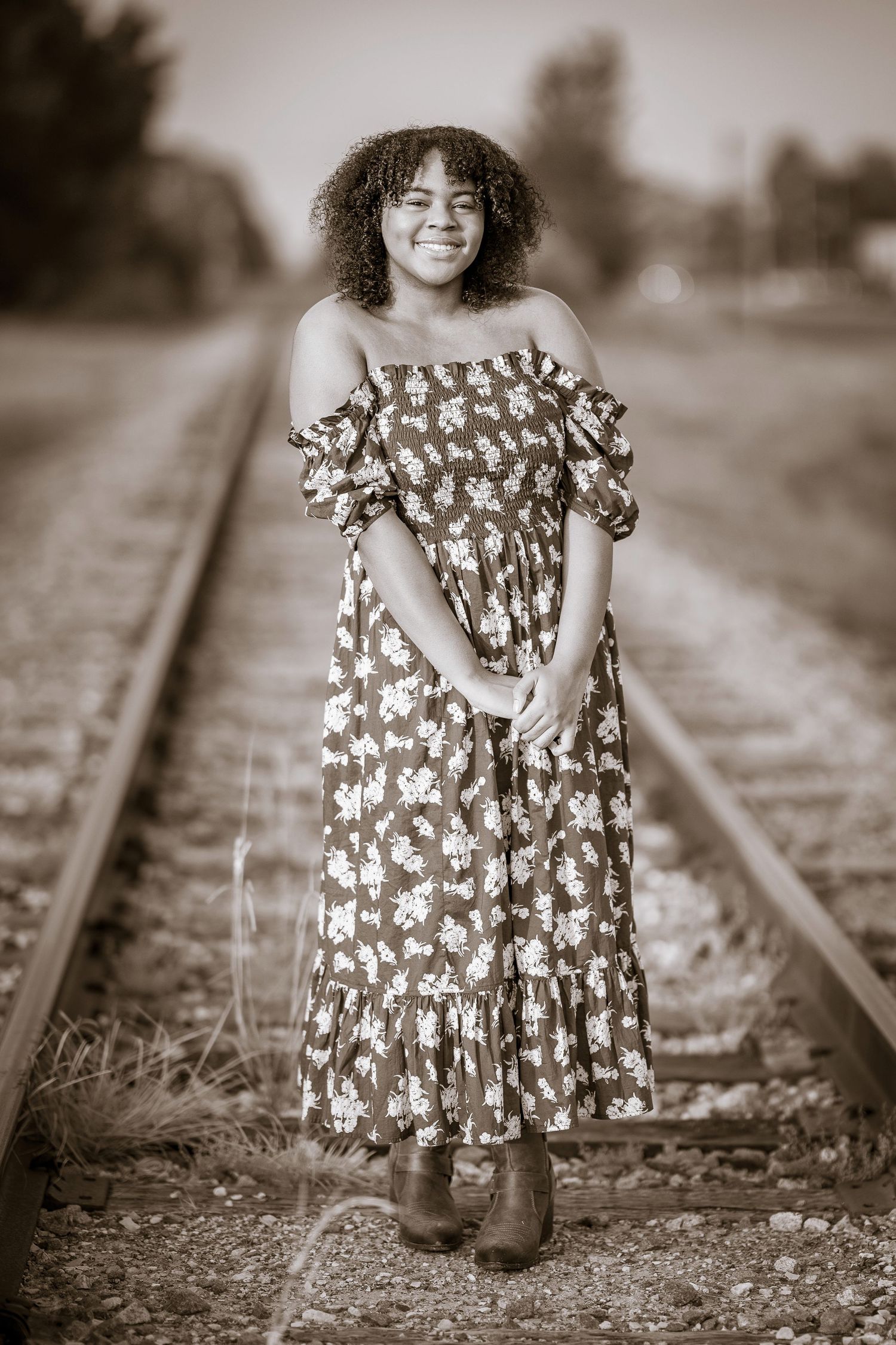 Sepia-toned portrait of a young Black woman in a floral dress standing on railroad tracks.