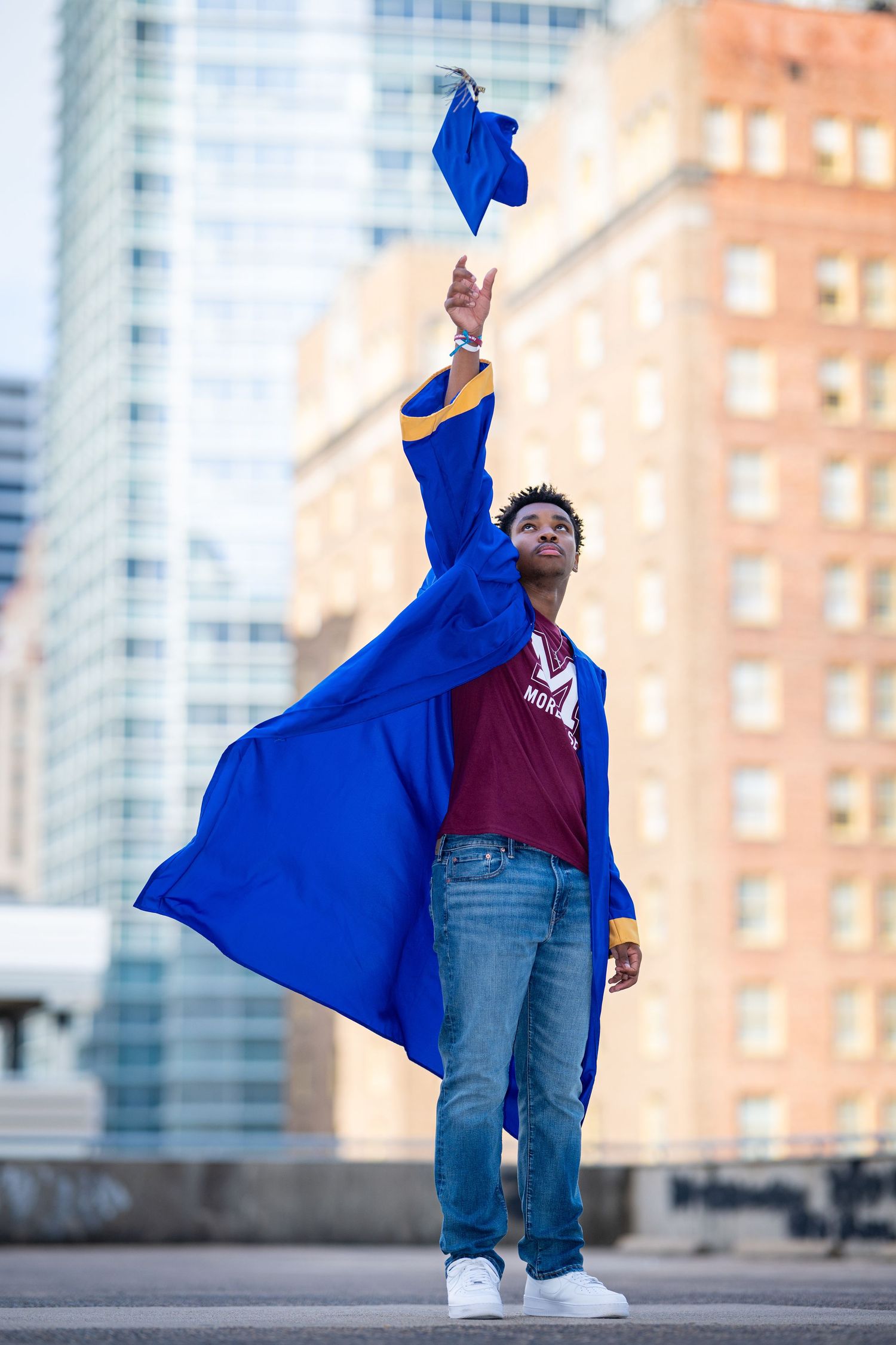 Young Black male graduate tossing blue cap in the air wearing graduation gown in the city.