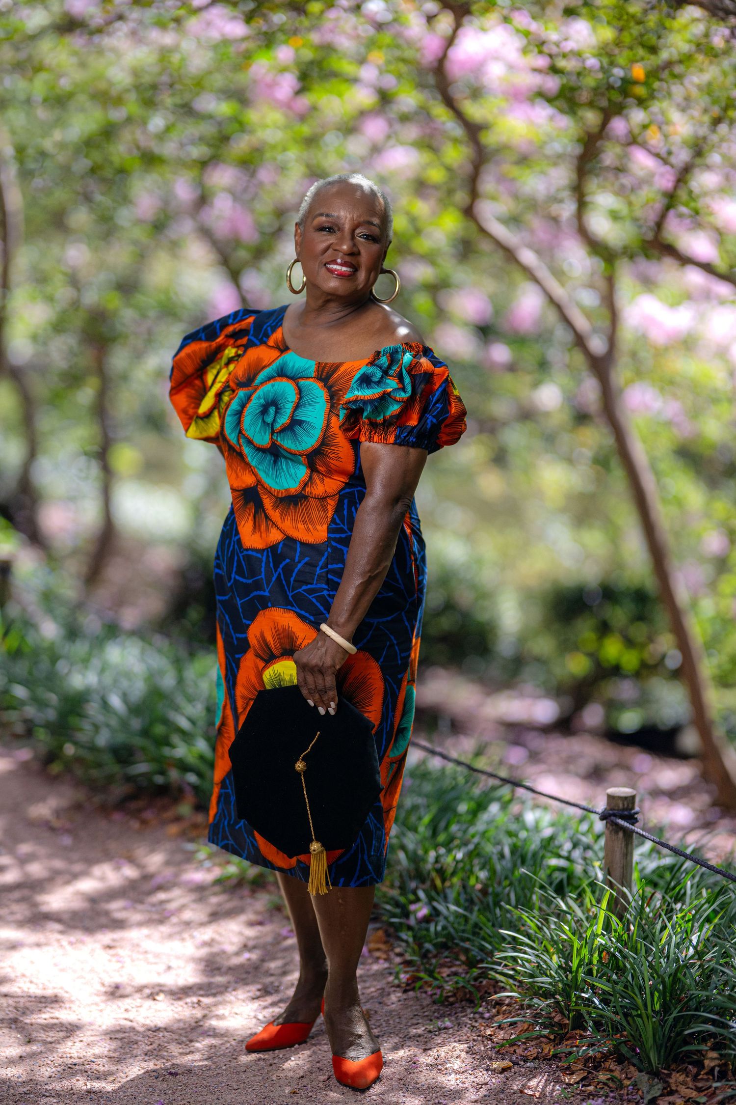 Woman in vibrant African print dress with floral details poses on garden path surrounded by blooming trees.