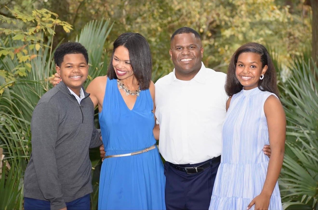 Happy family of four smiling together outdoors, wearing blue and white outfits among lush green foliage.