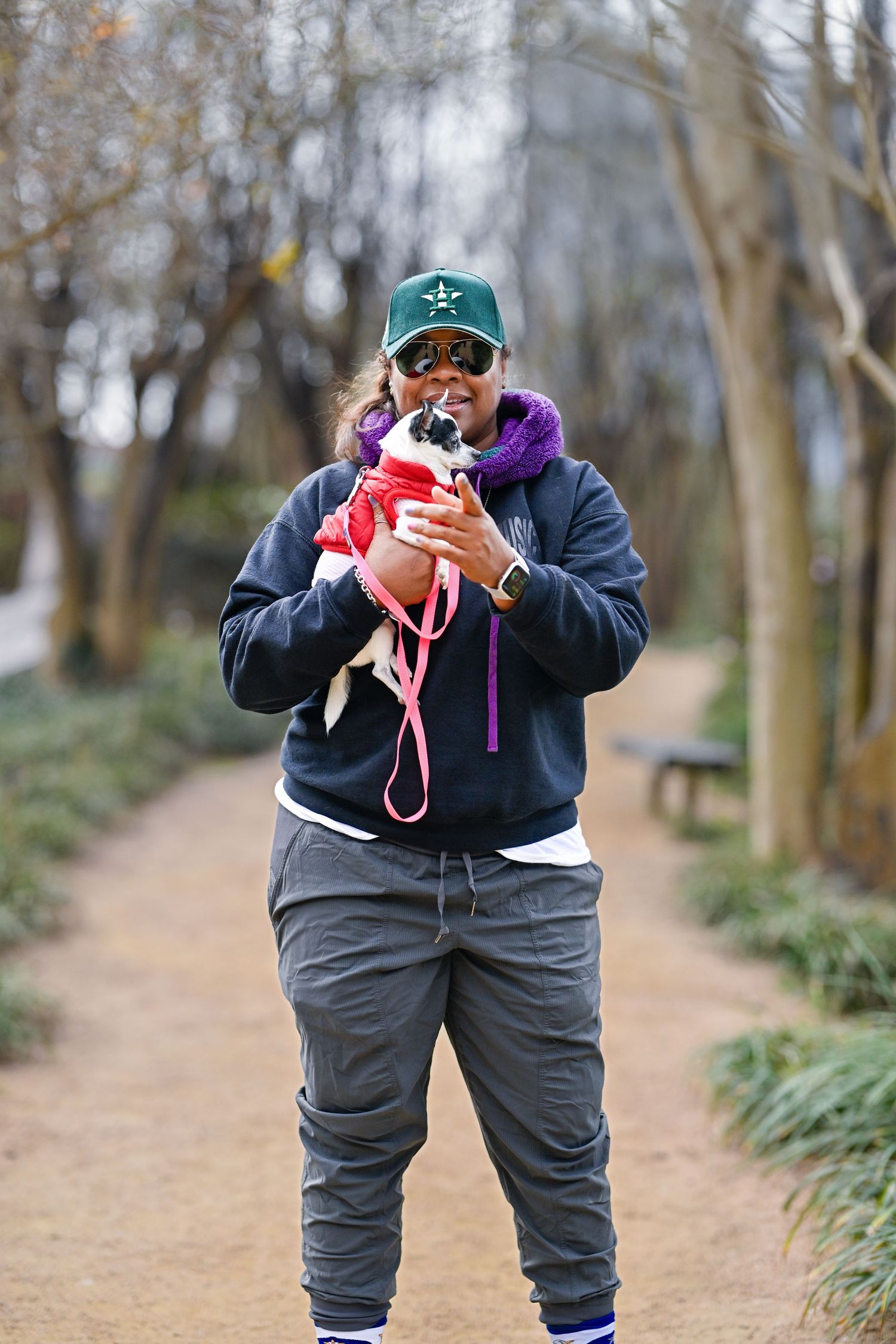 Person in navy hoodie and green cap kissing small dog in red jacket on park path.