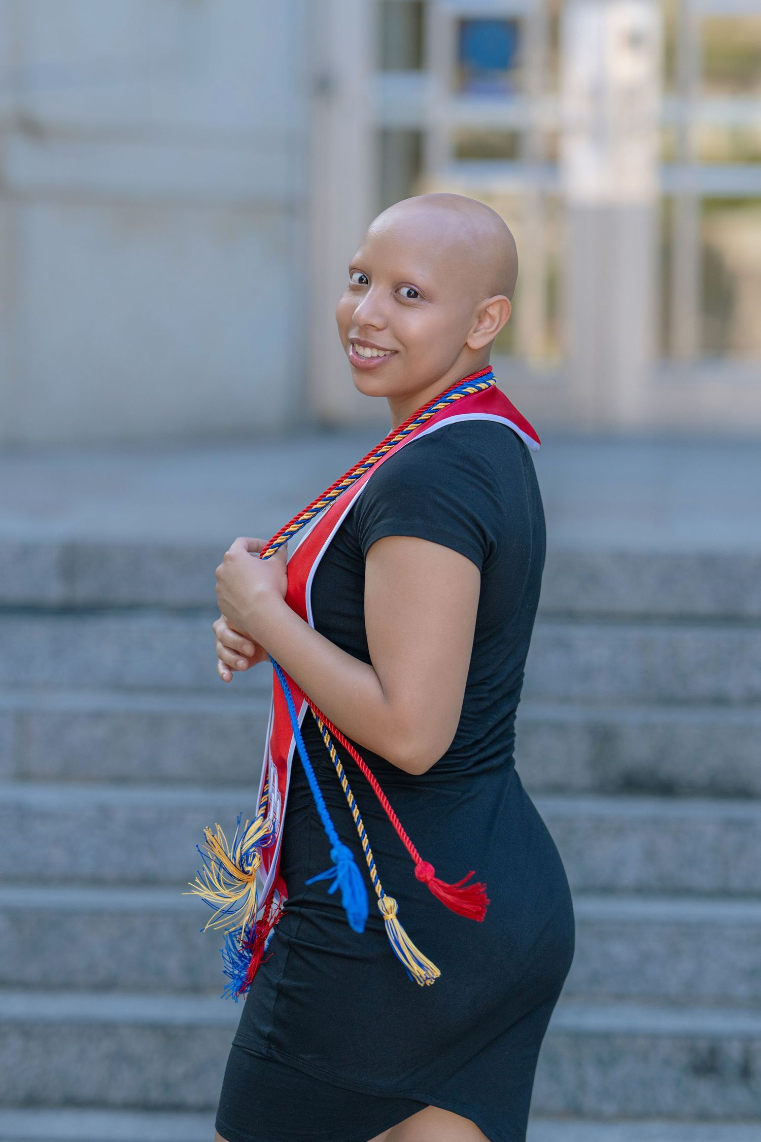 Smiling bald woman in a black dress with colorful graduation cords posing on outdoor steps.