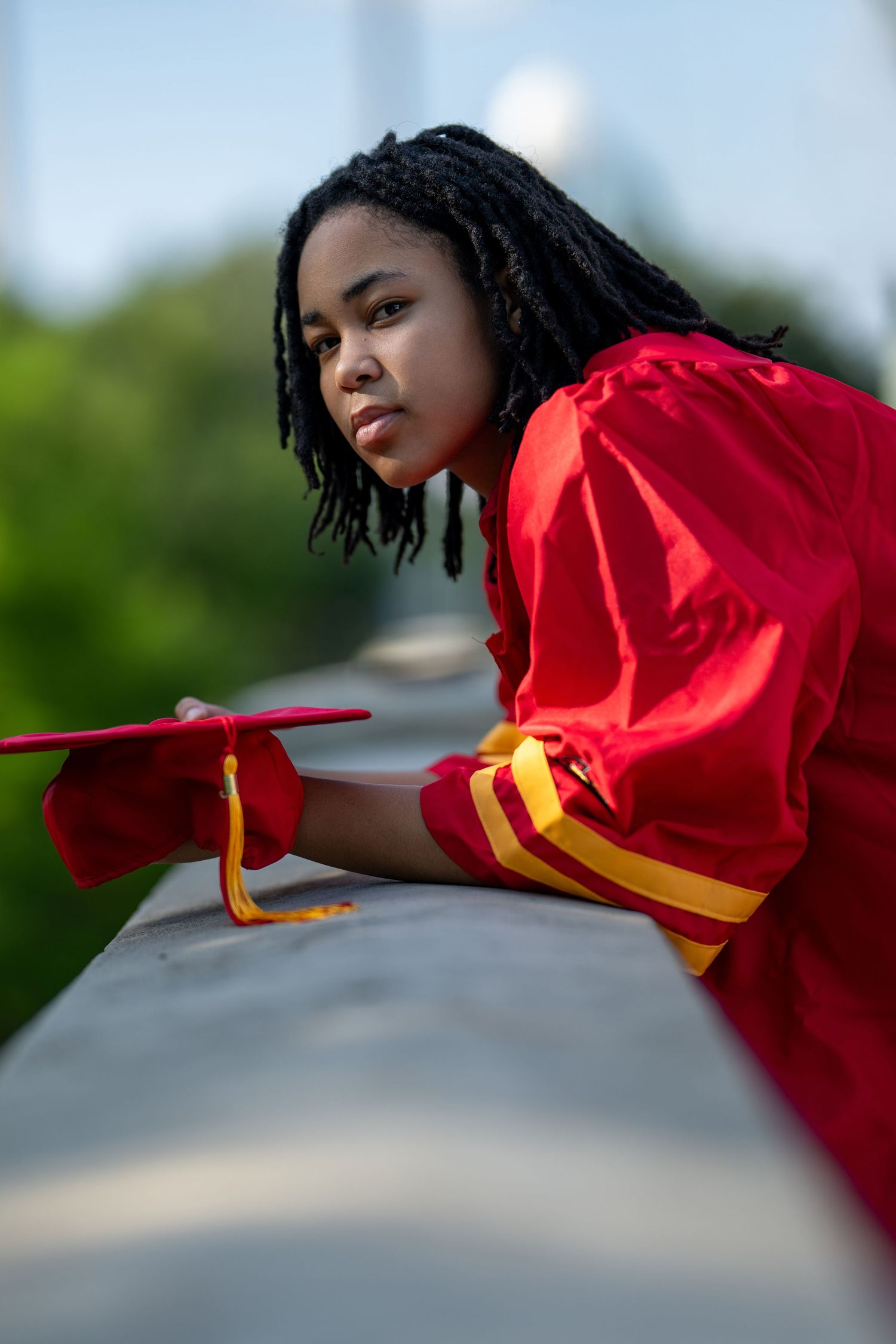 Young Black woman in red graduation gown holding cap while leaning on a ledge outdoors.