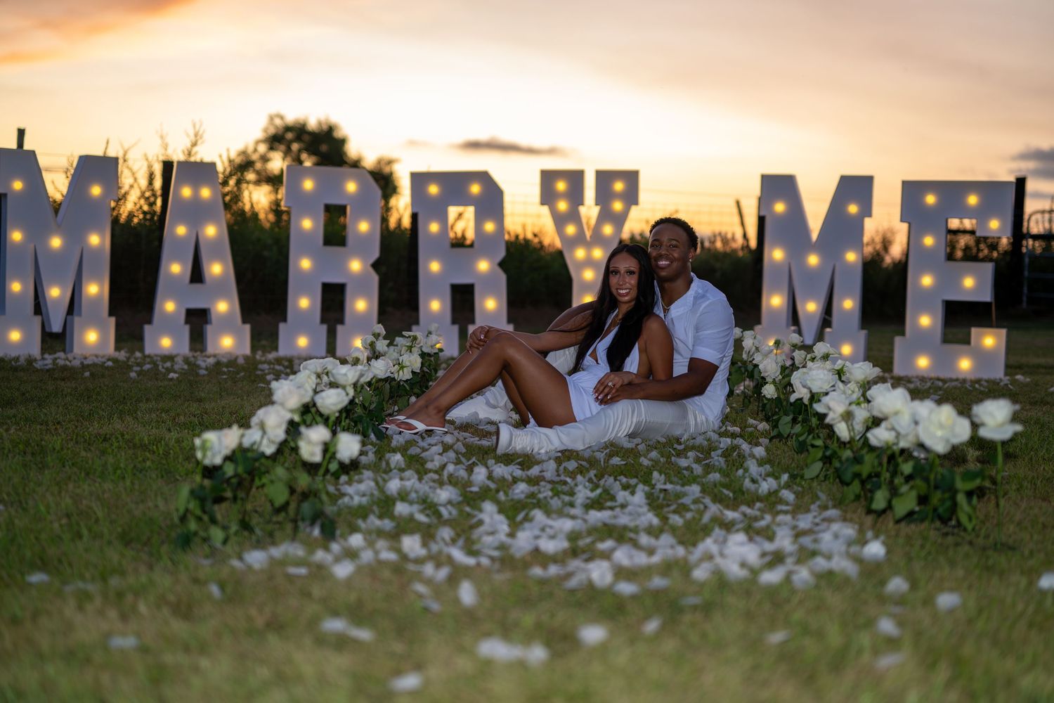 Couple sitting together in front of illuminated Marry Me marquee letters at sunset with white roses.