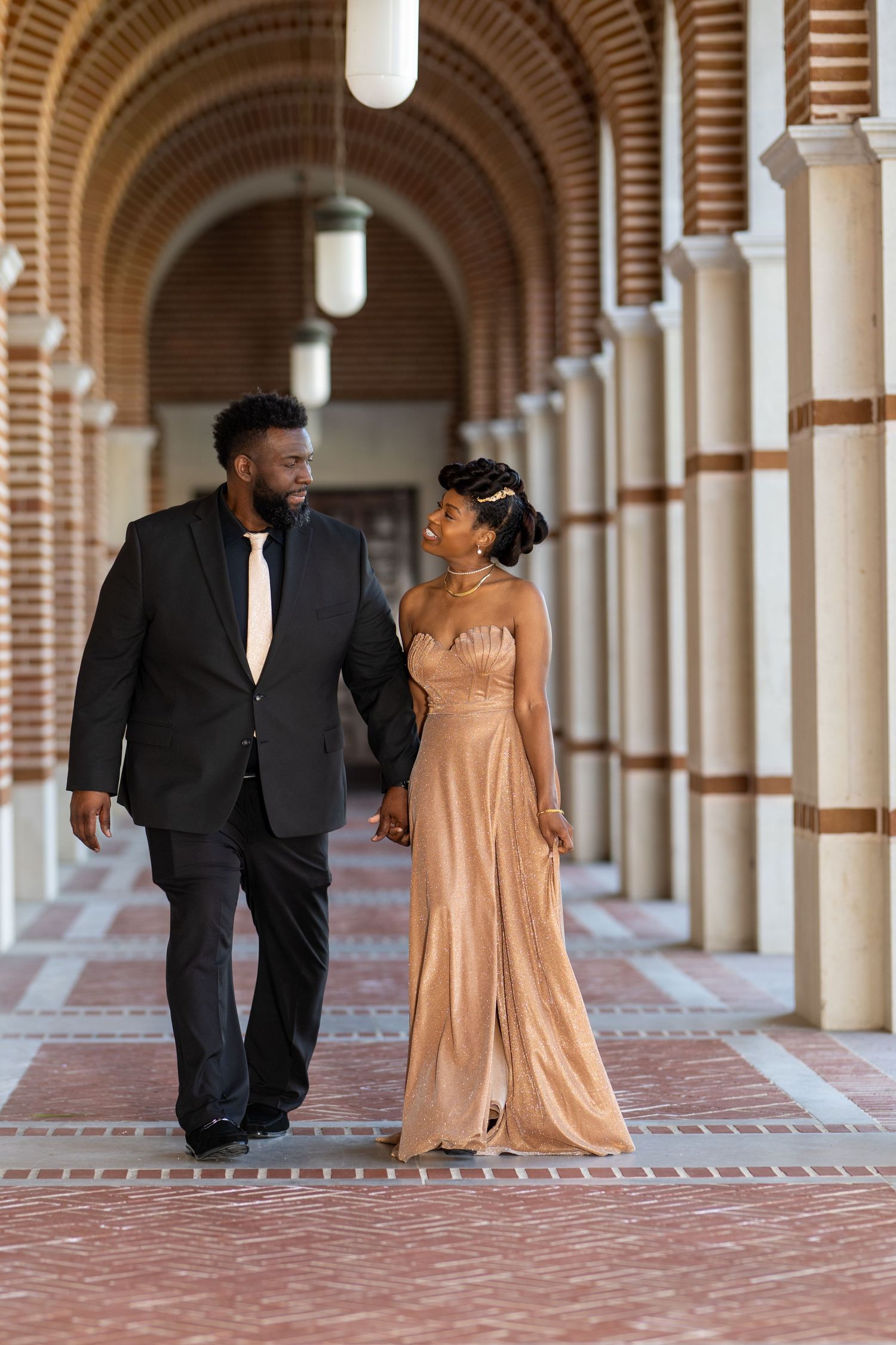 A couple in formal attire walks hand in hand through an elegant arched brick corridor.