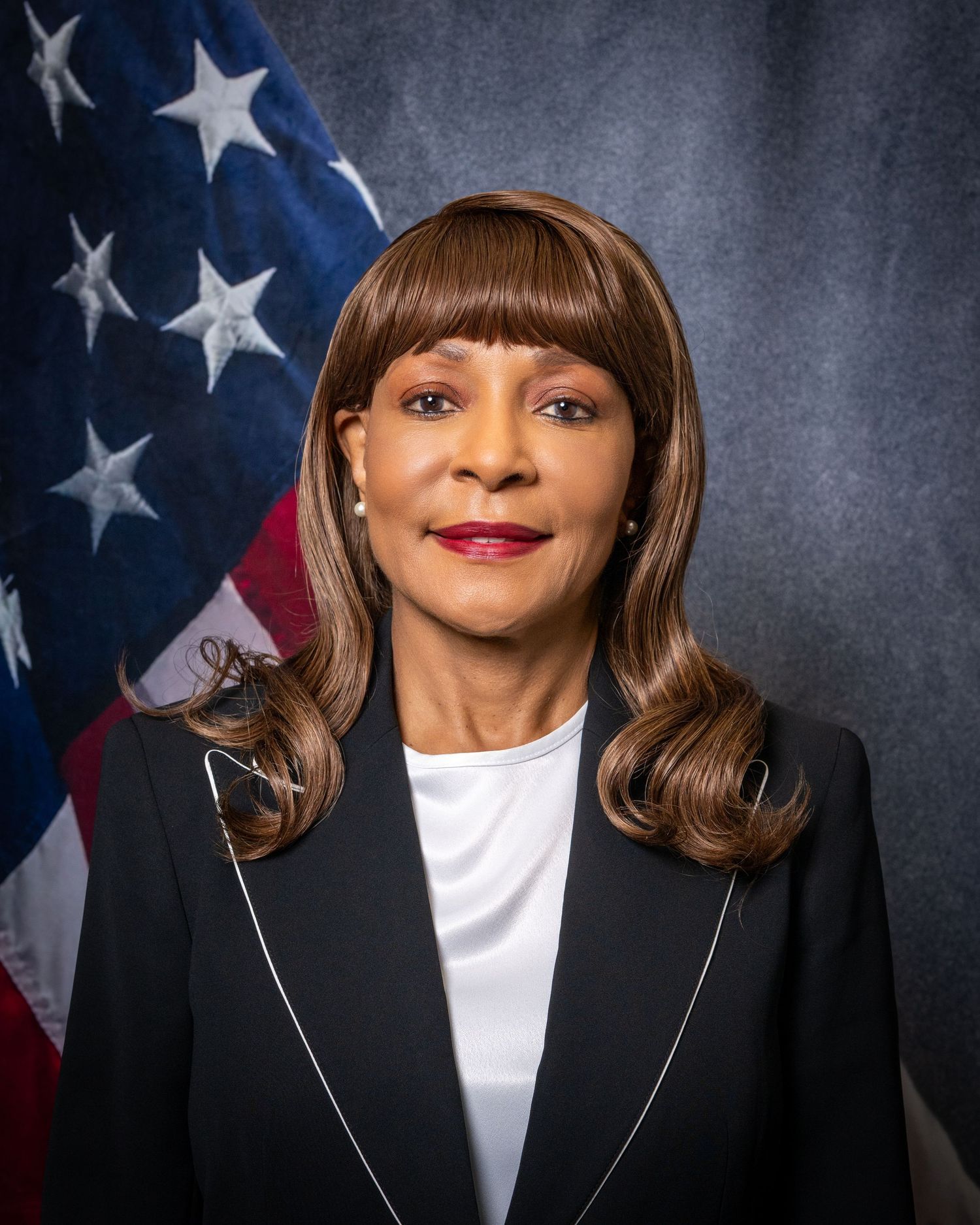 Professional headshot of an elegant woman in a black blazer with styled brown hair before an American flag.