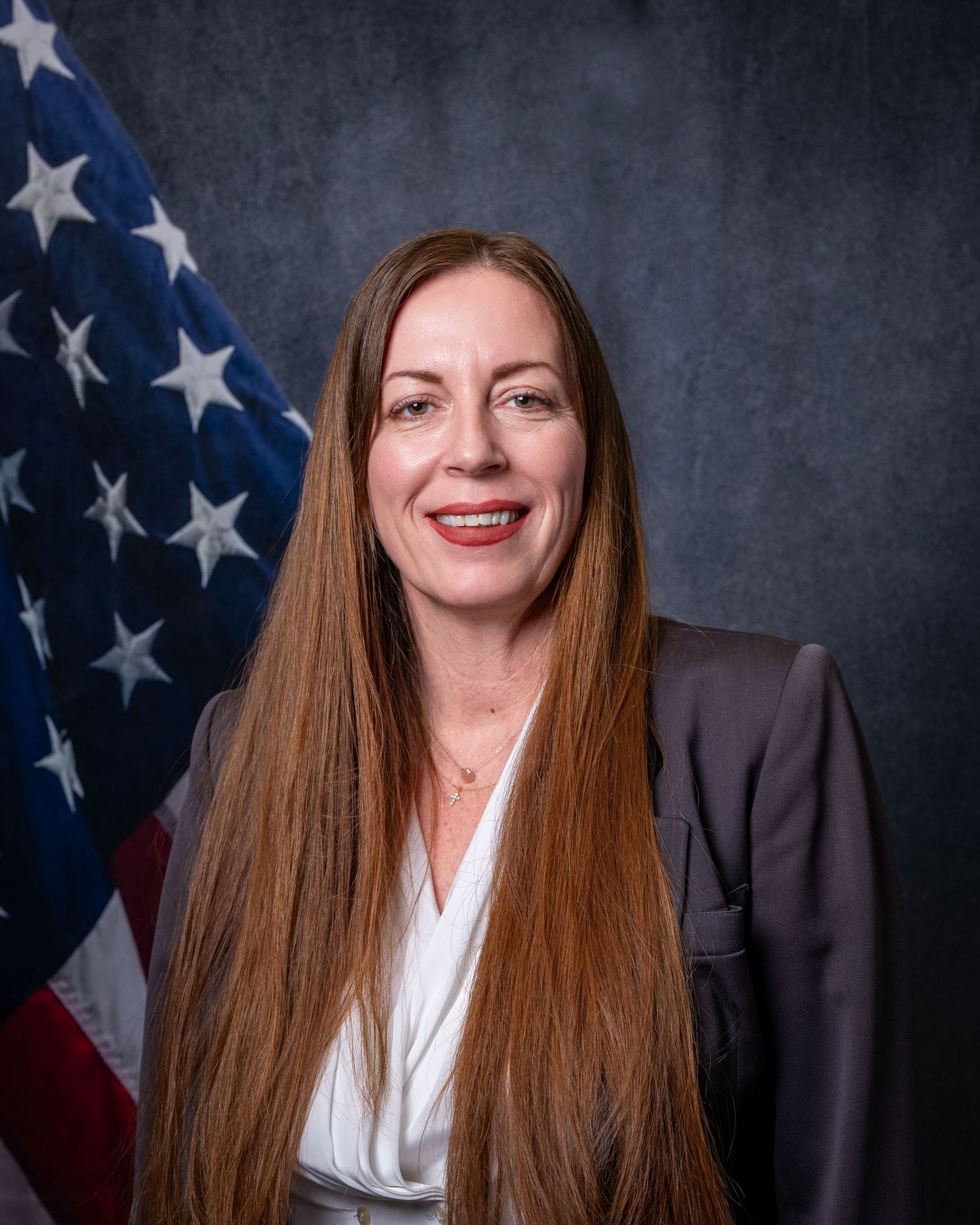 Professional portrait of a smiling woman with long auburn hair in a gray blazer before an American flag.