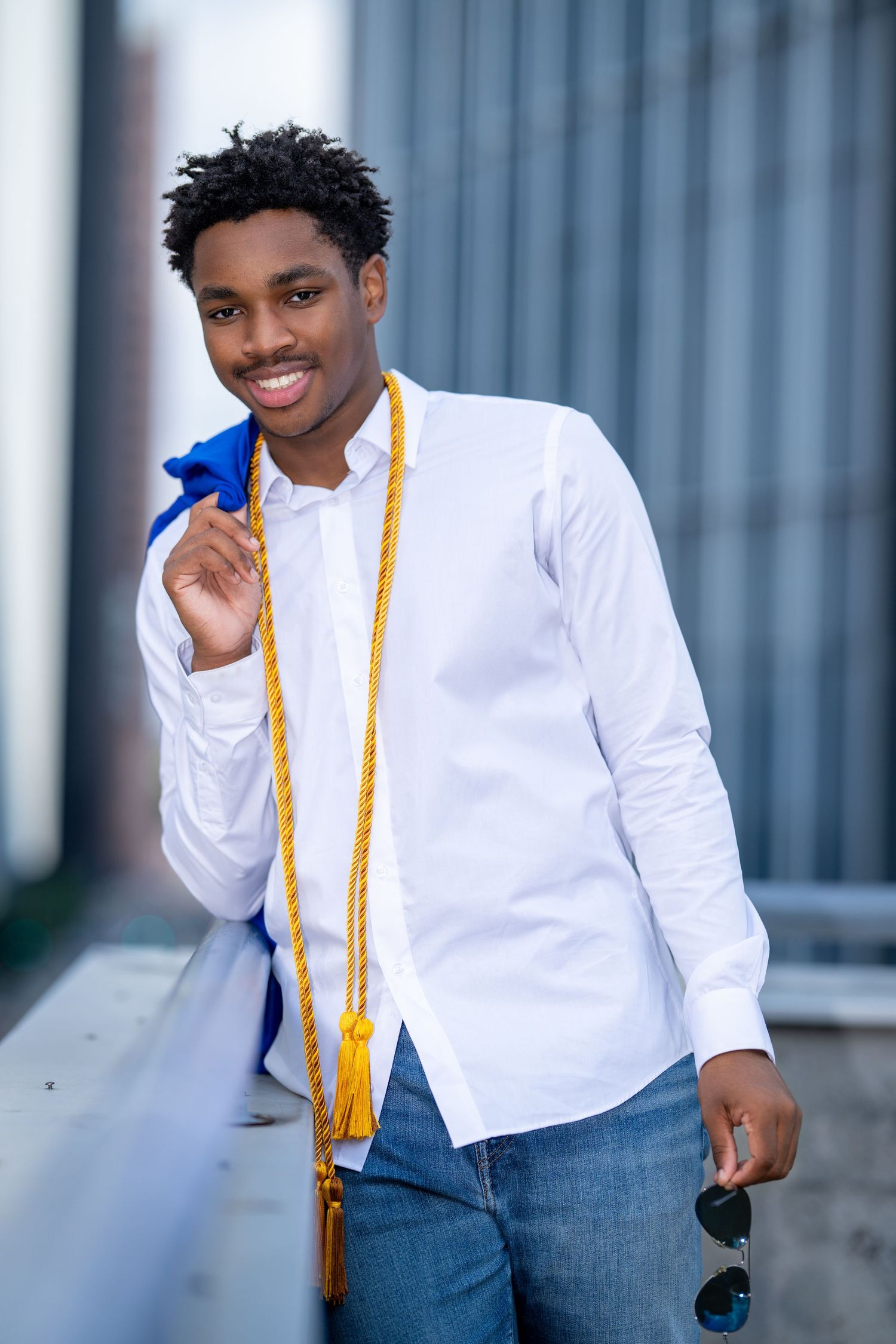 Smiling young Black male graduate in white shirt holding blue gown and gold honor cords.