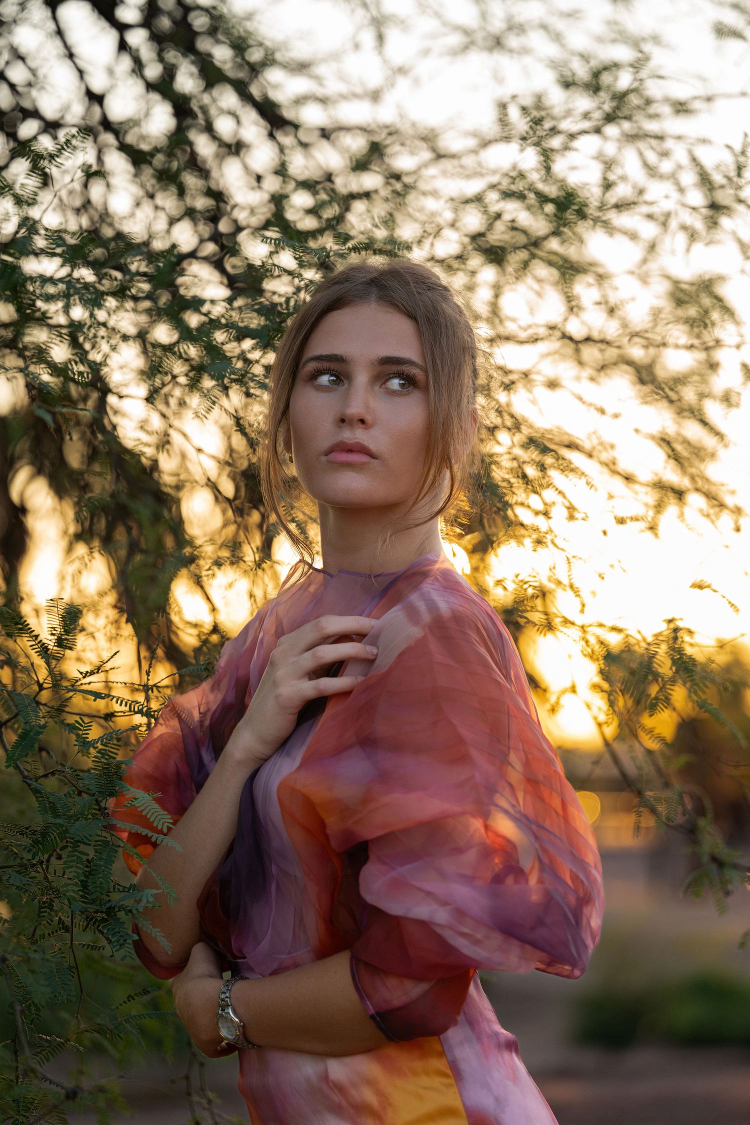 A young woman in a multicolored sheer top gazes upward while standing near green foliage at sunset.