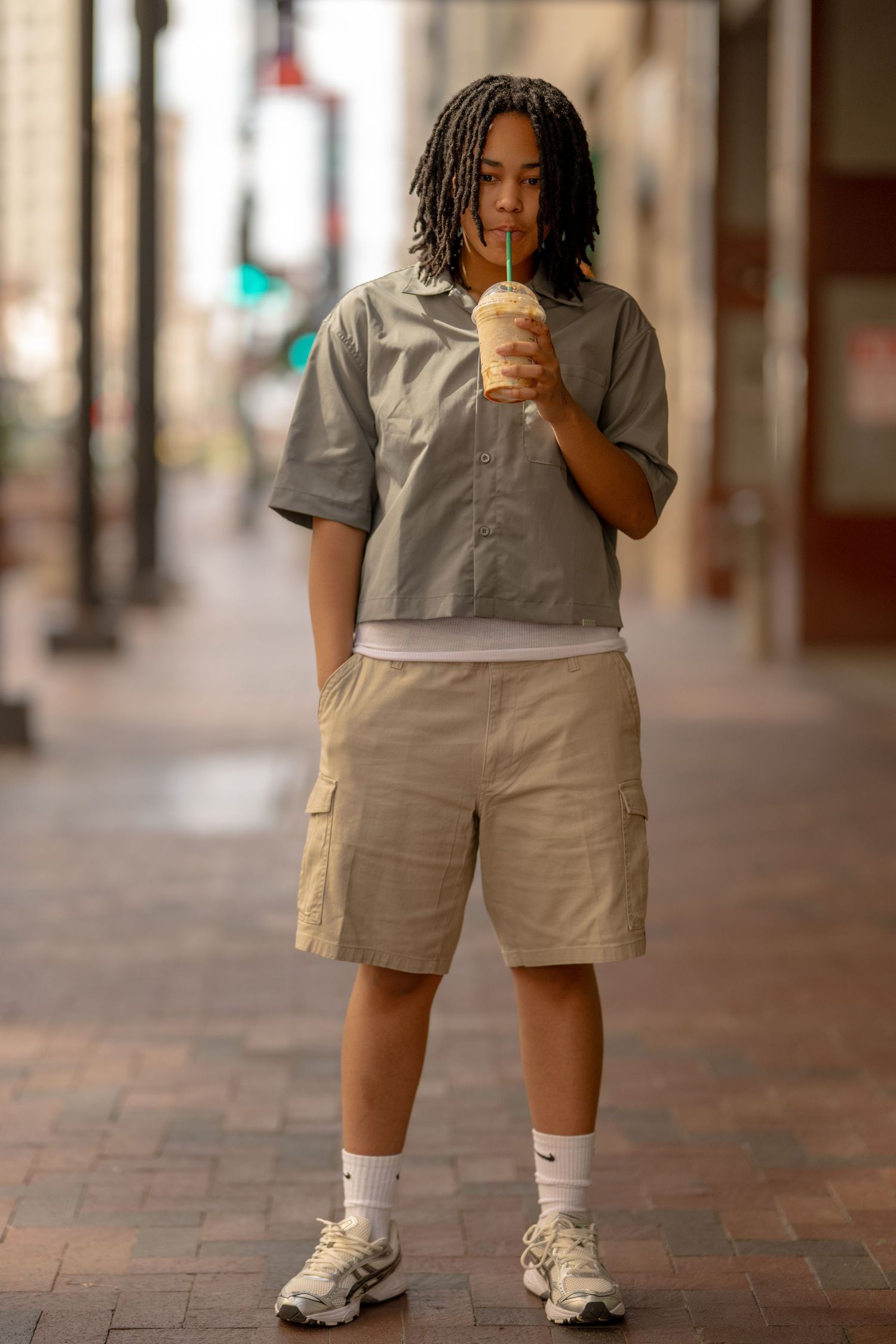 Young person with locs sipping an iced drink while standing on an urban sidewalk in casual attire.
