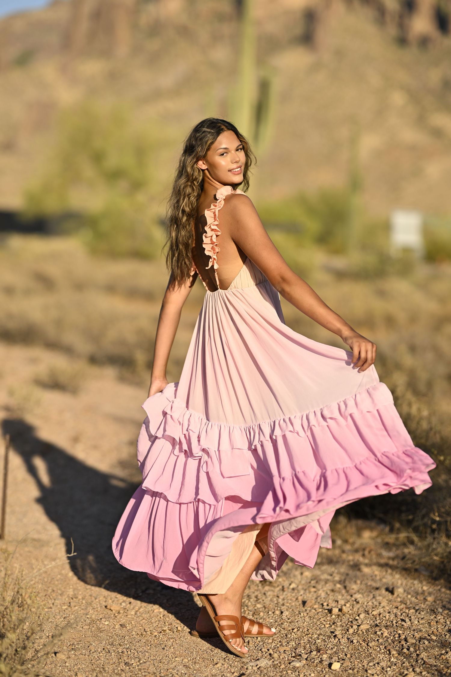 Woman in flowing pink ruffled maxi dress twirling on desert path with saguaro cactus in background.
