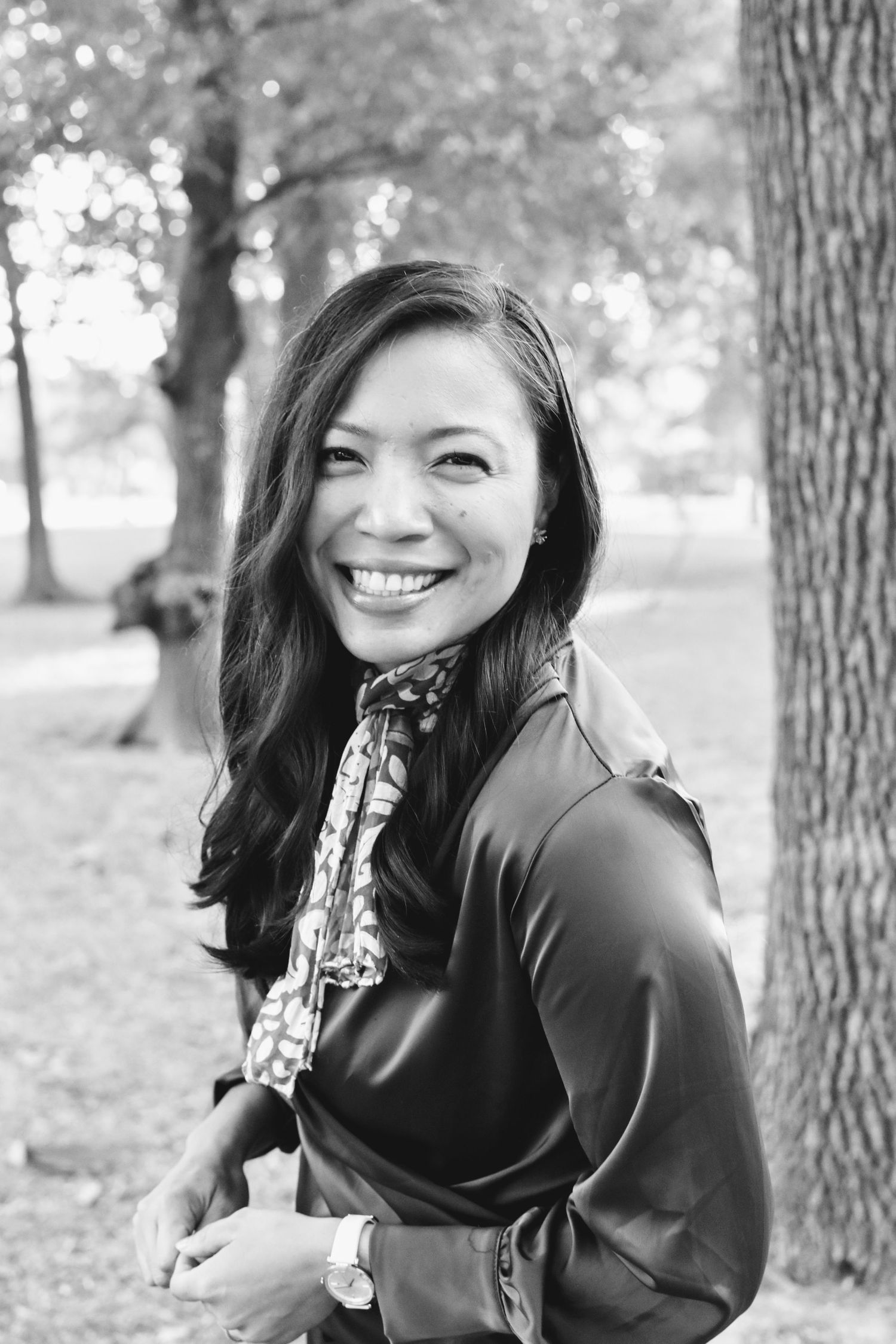 Smiling woman with long dark hair wearing a scarf poses outdoors near trees in black and white photo.