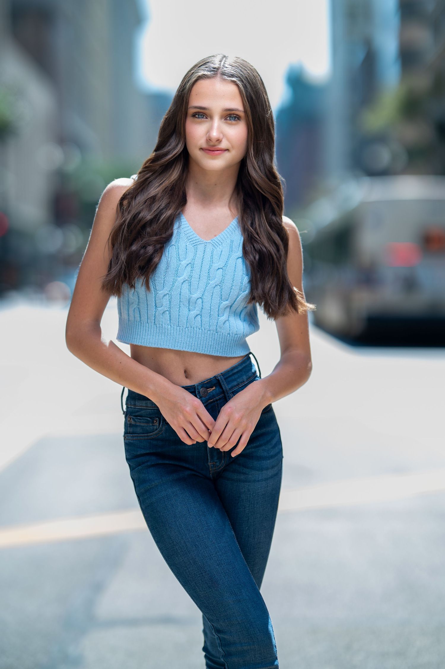 Teen girl with long brown hair in a blue crop top and jeans poses on a city street.