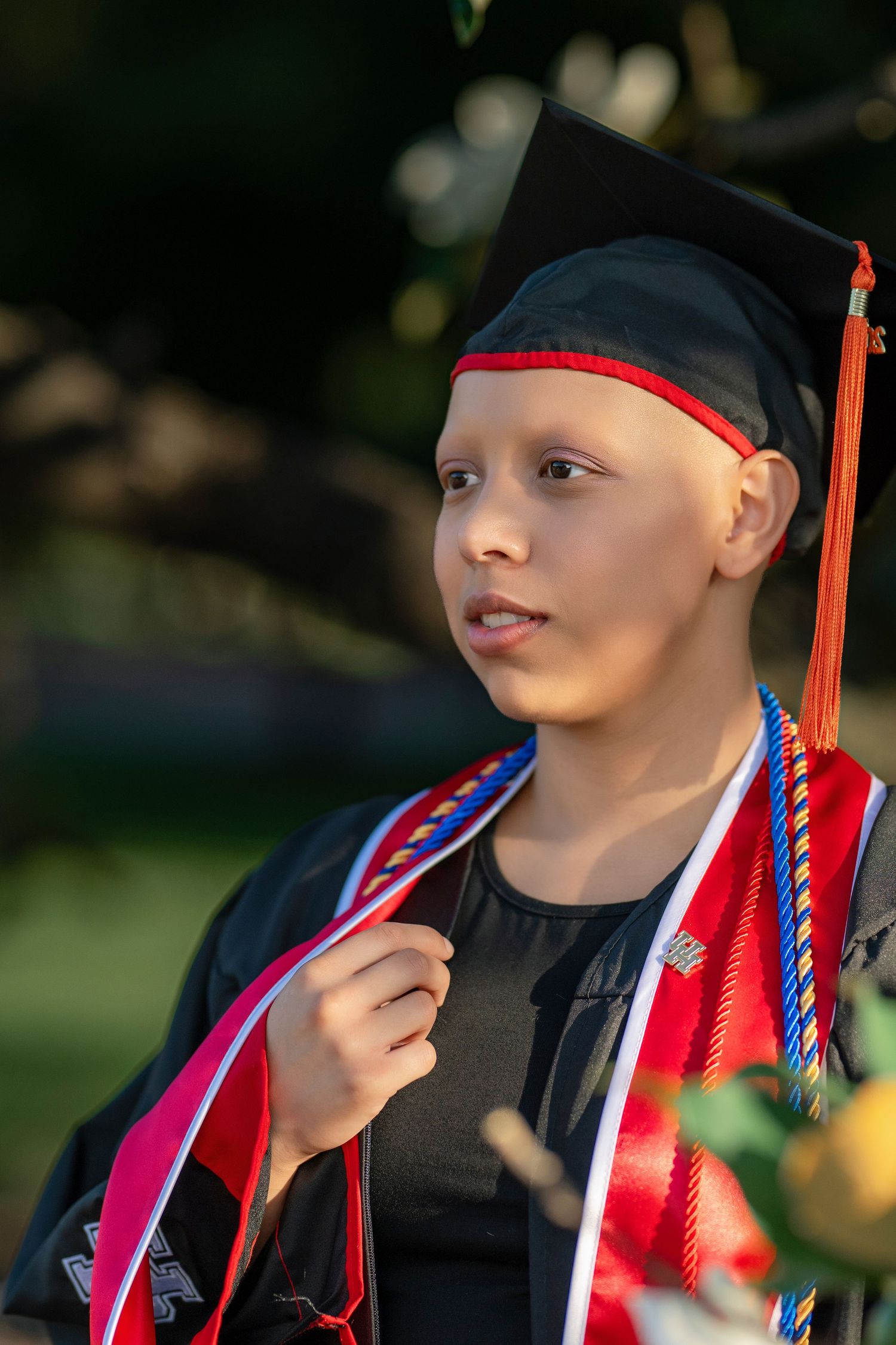 Young student in black graduation cap and gown with red and blue honor cords outdoors.