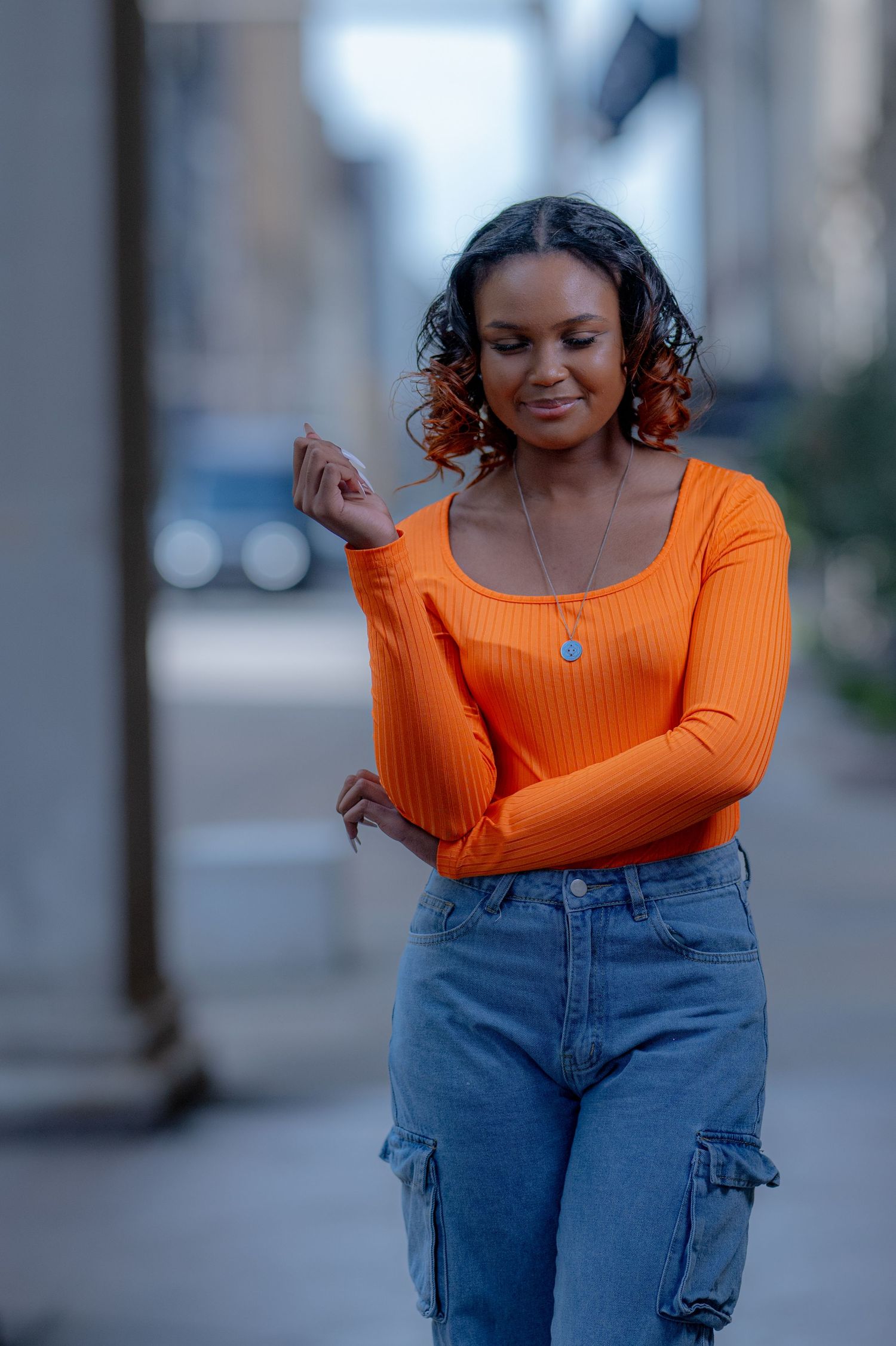 Young woman in orange ribbed top and cargo jeans walking on urban city street.