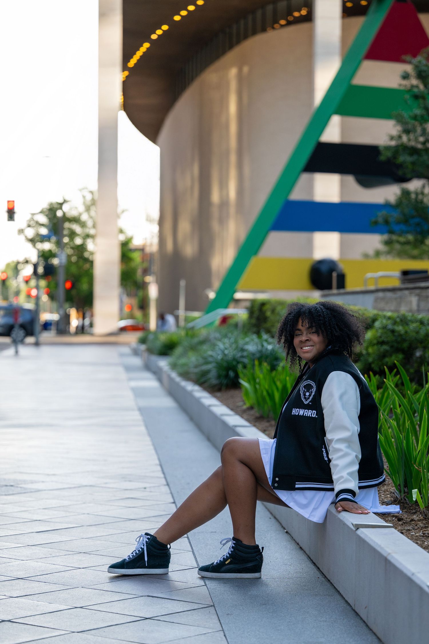 Young woman in varsity jacket and sneakers smiling while sitting on urban plaza ledge near colorful sculpture.