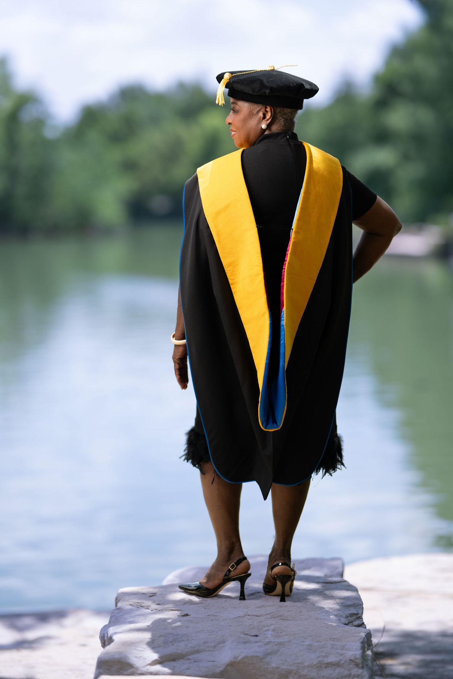 Woman in black graduation gown and cap with gold hood stands on rock by lake.