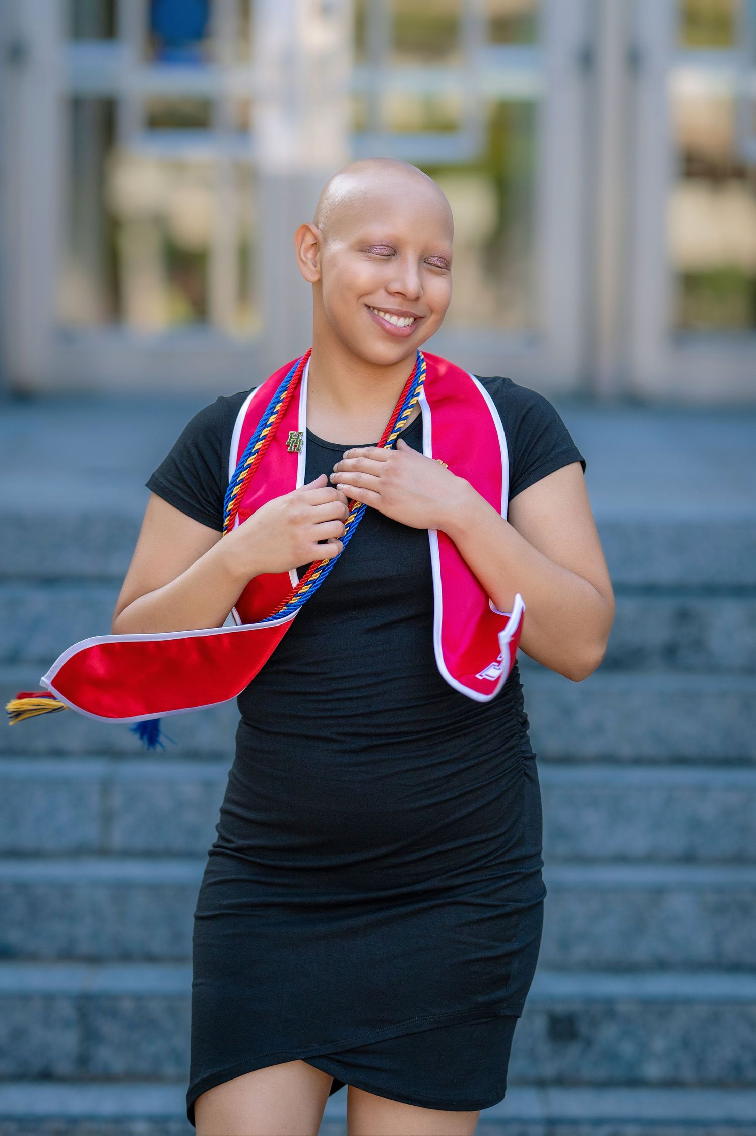 Smiling bald woman in black dress with pink graduation stole standing on outdoor steps.