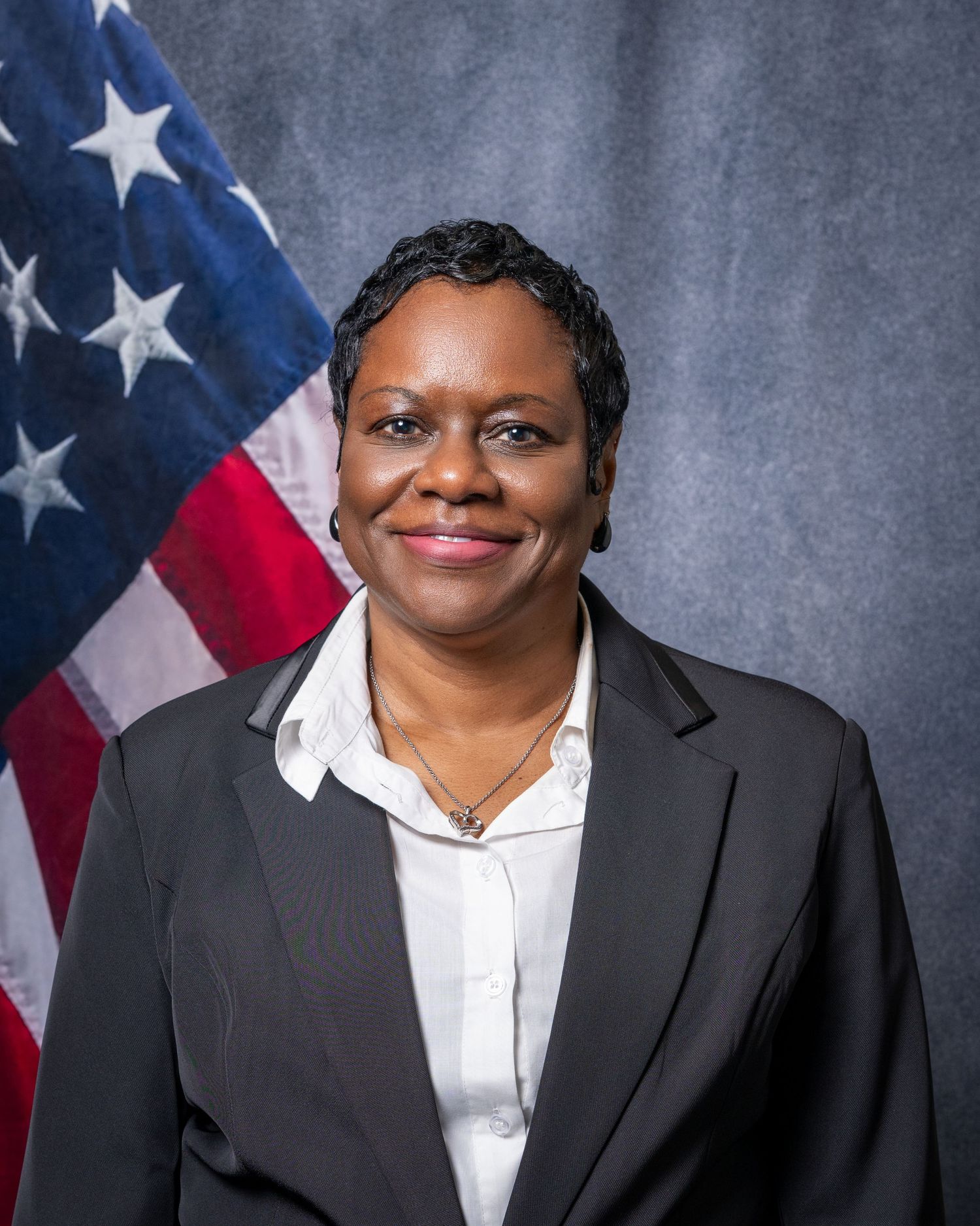 Professional headshot of a woman in a black blazer with white blouse before an American flag.