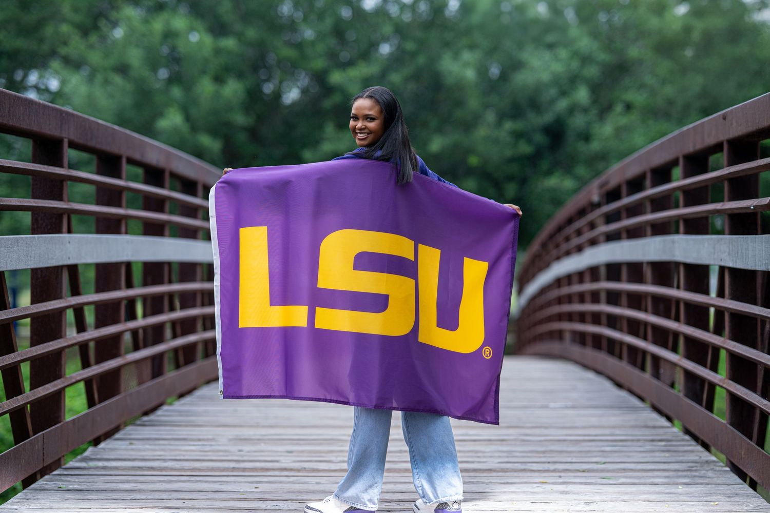 Young woman holding a purple LSU flag on a wooden bridge surrounded by green trees.