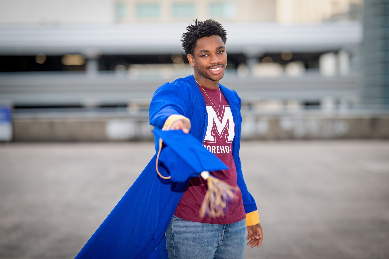 Happy young Black male graduate extending hand wearing blue graduation gown and Morehouse shirt.