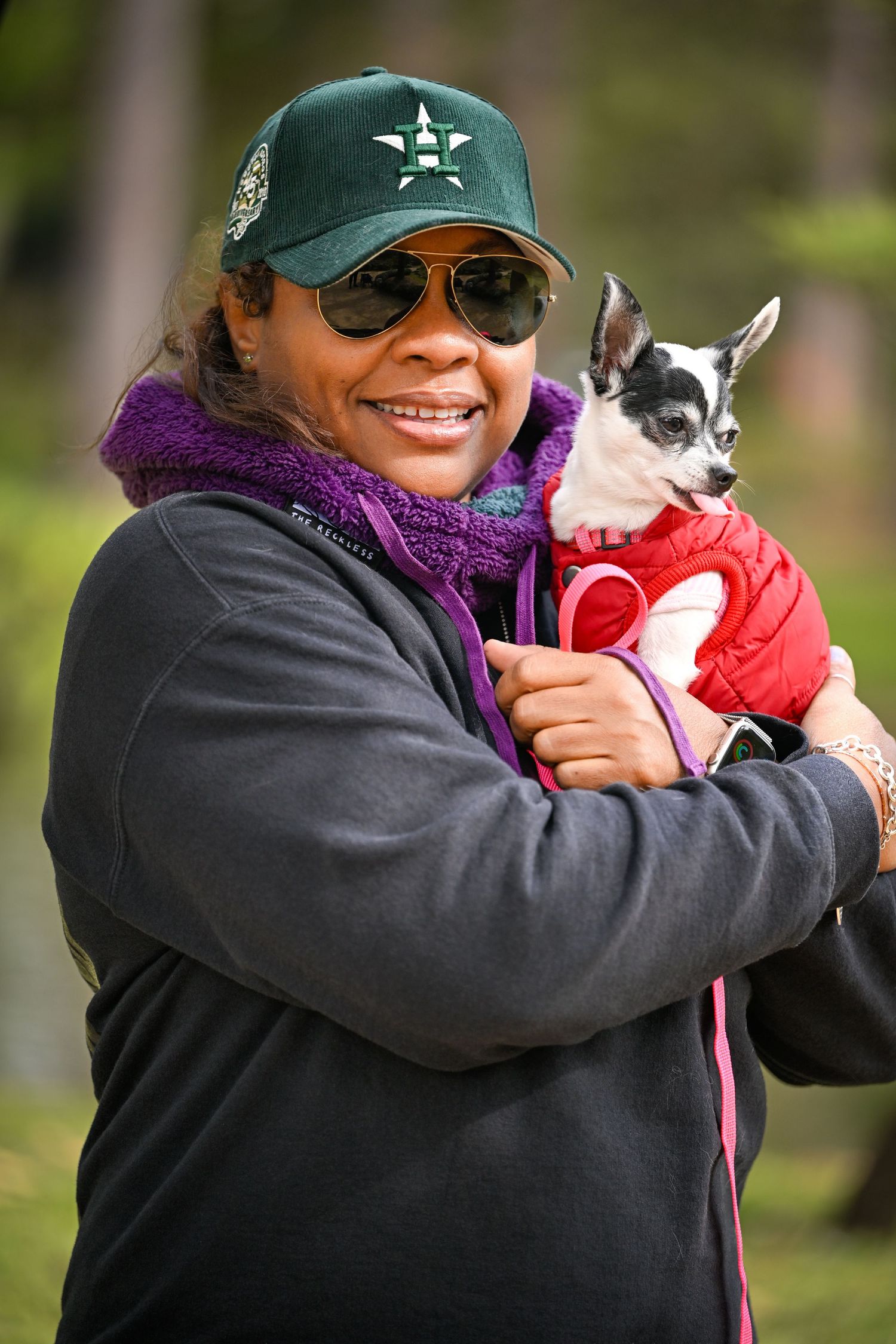 Smiling woman in green baseball cap holding small dog in red jacket outdoors.