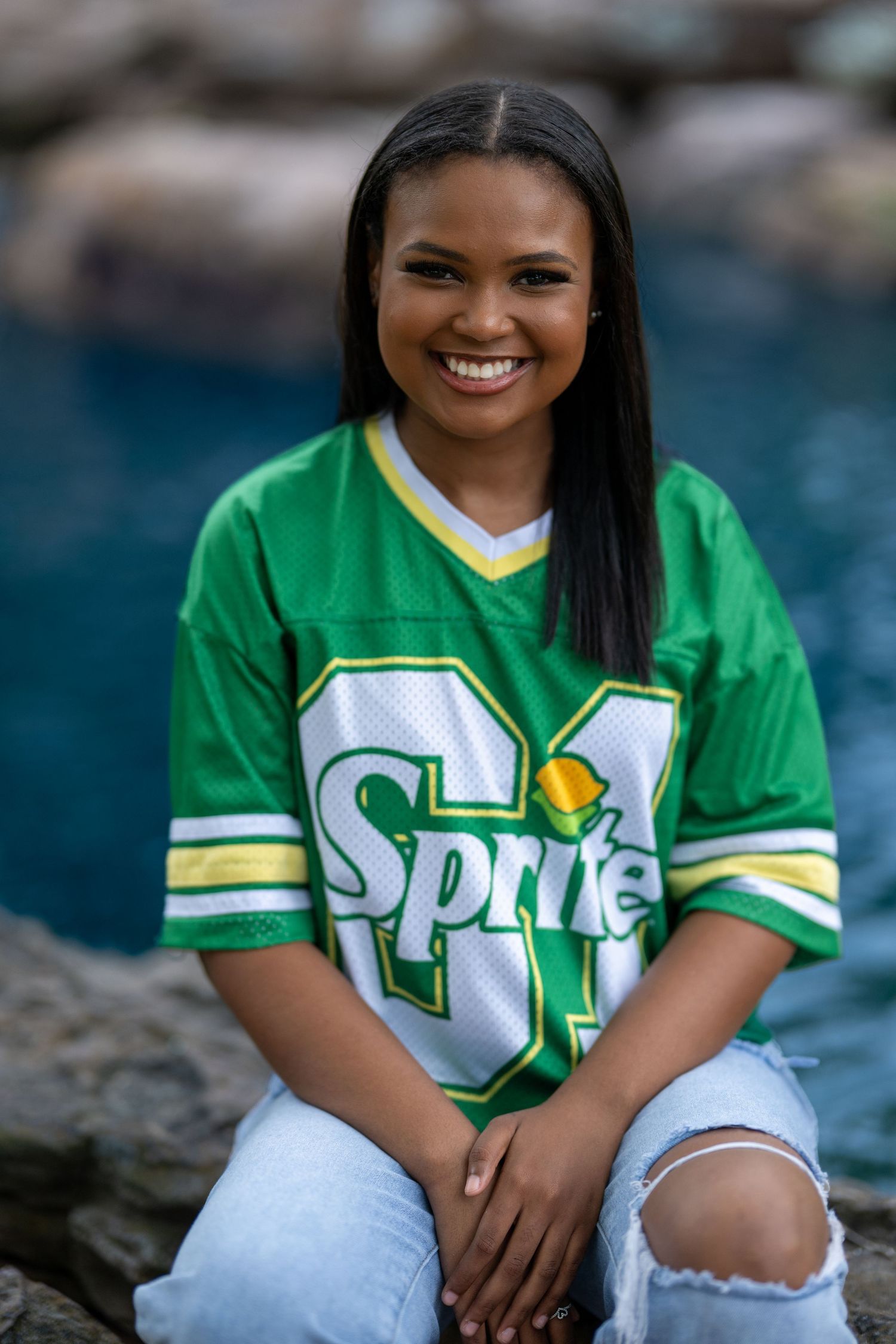 Young woman smiling in a green Sprite football jersey and ripped jeans, sitting on rocks near water.