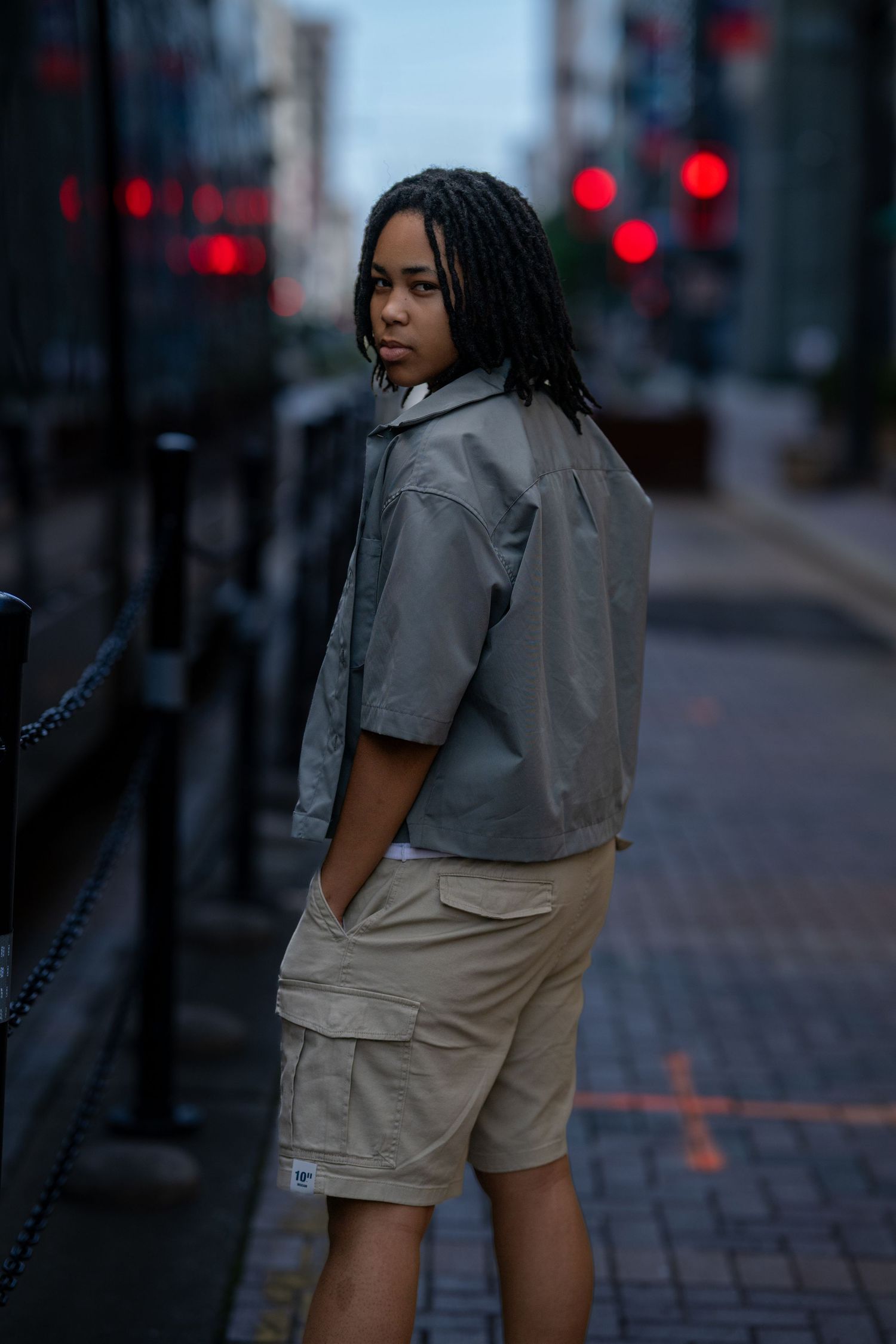 Young person with braids in a gray jacket and khaki cargo shorts standing on a city sidewalk at dusk.