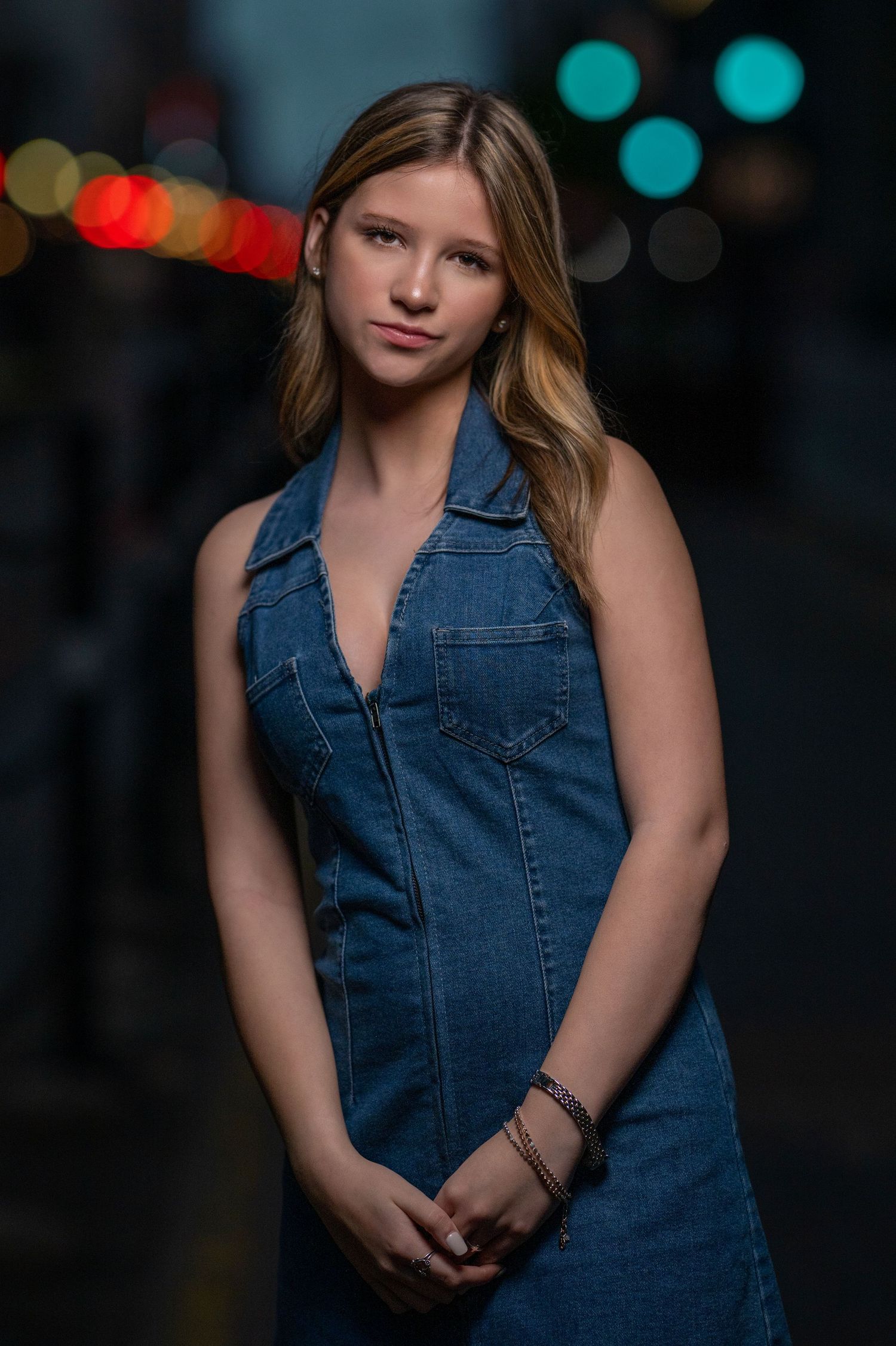 Teen girl in sleeveless denim dress posing confidently on a city street at night.