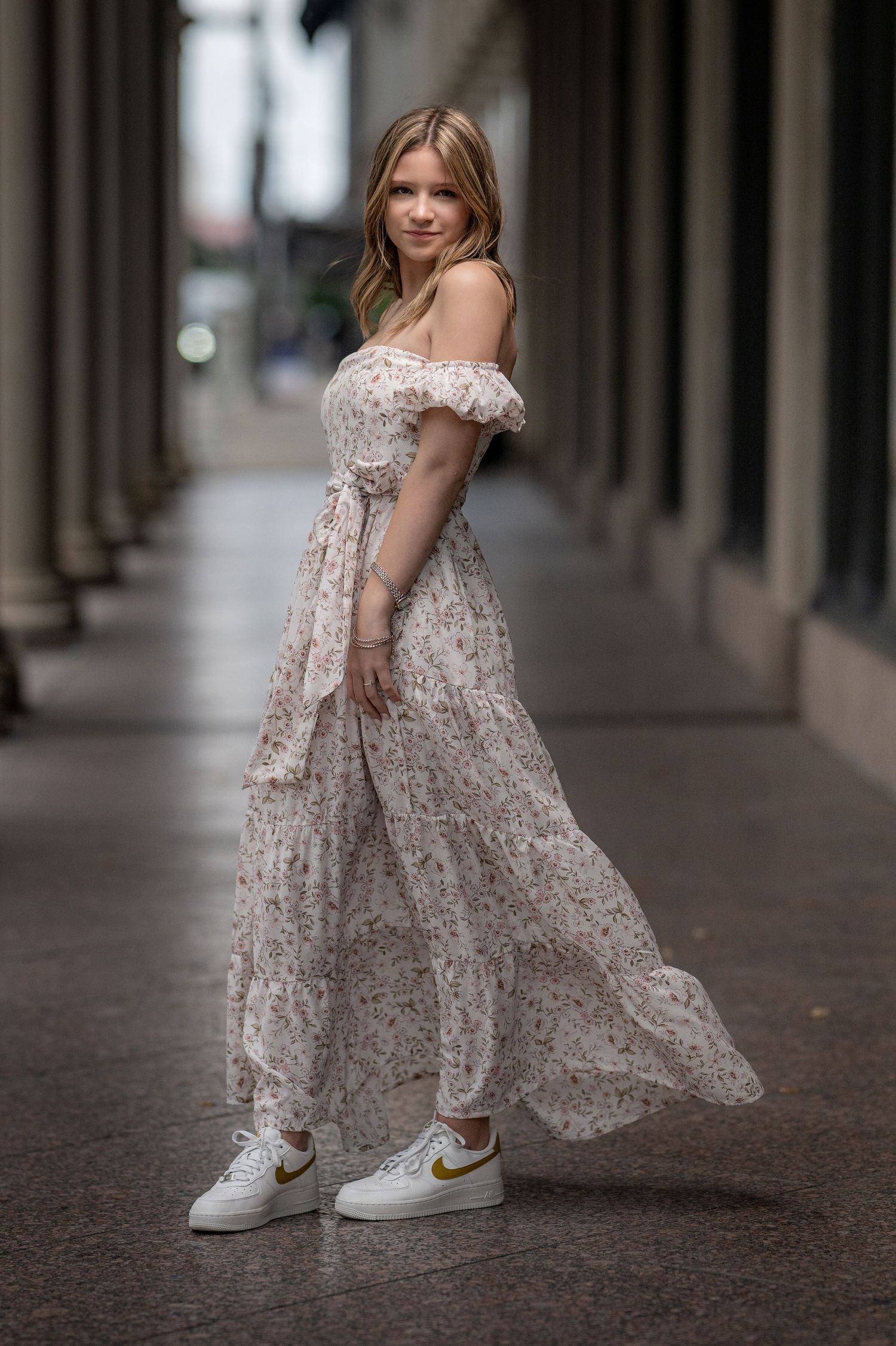 Blonde girl in a floral off-shoulder maxi dress and white Nike sneakers posing in an urban corridor.