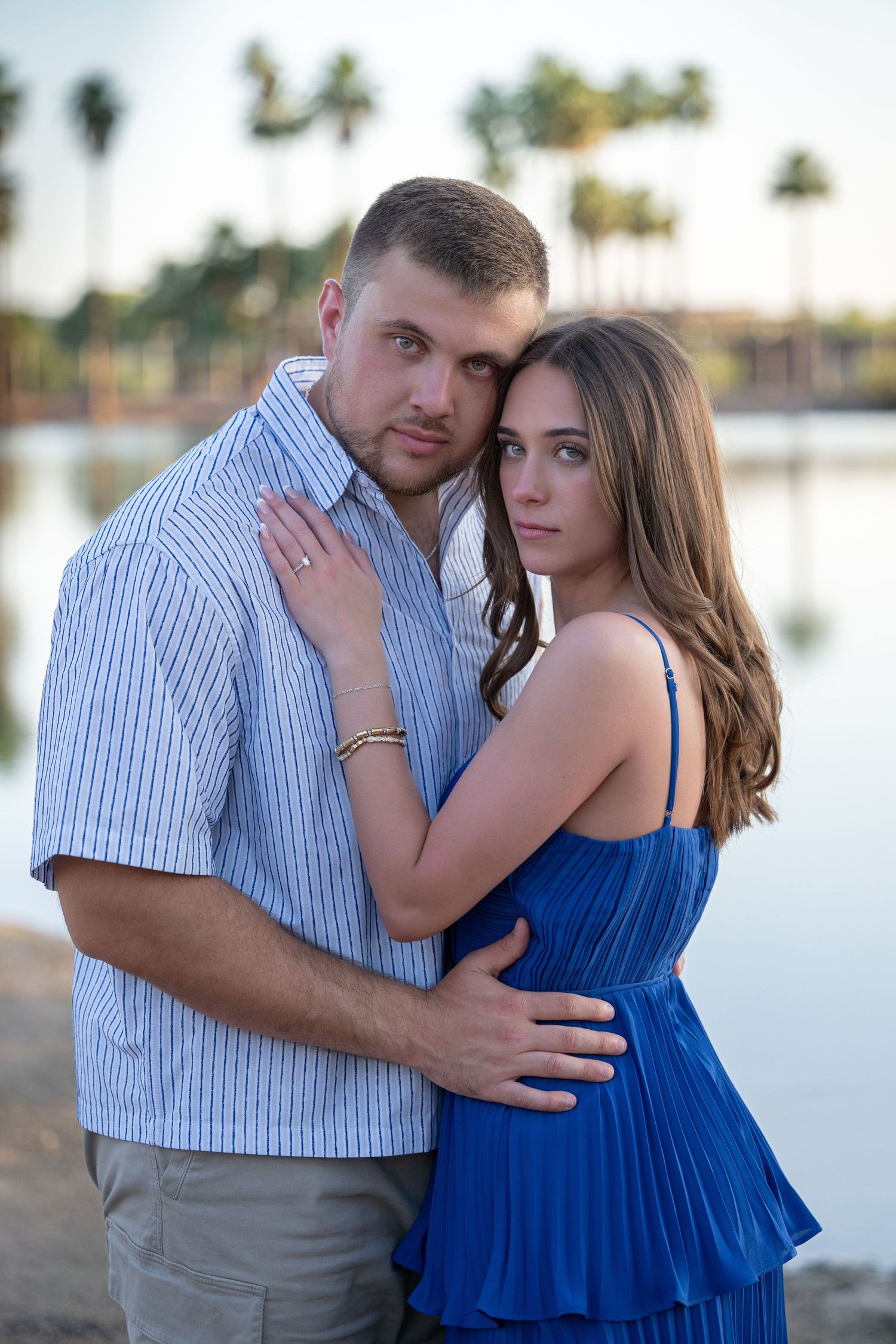 A young couple embraces near a reflective lake with palm trees in the background at dusk.