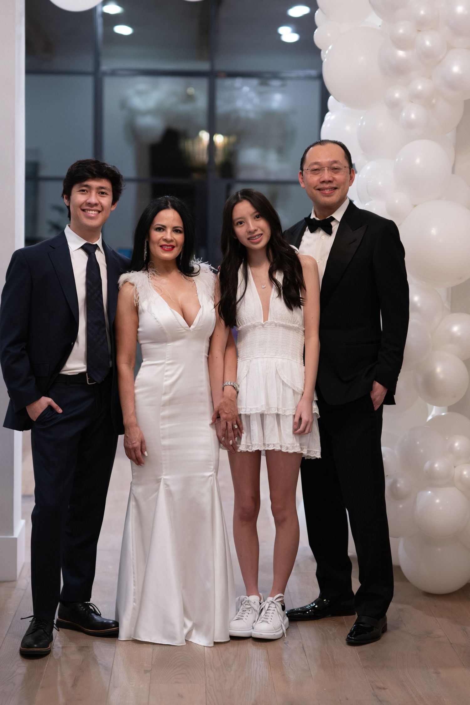 Family of four posing in formal attire with white balloon arch at elegant event.