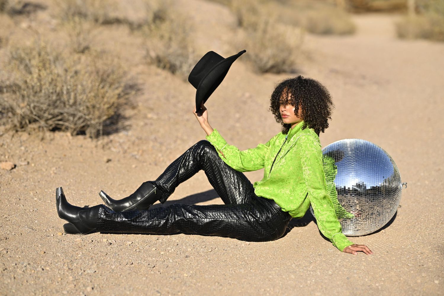 Stylish woman in green jacket and black patent boots sitting in desert sand holding cowboy hat beside disco ball.