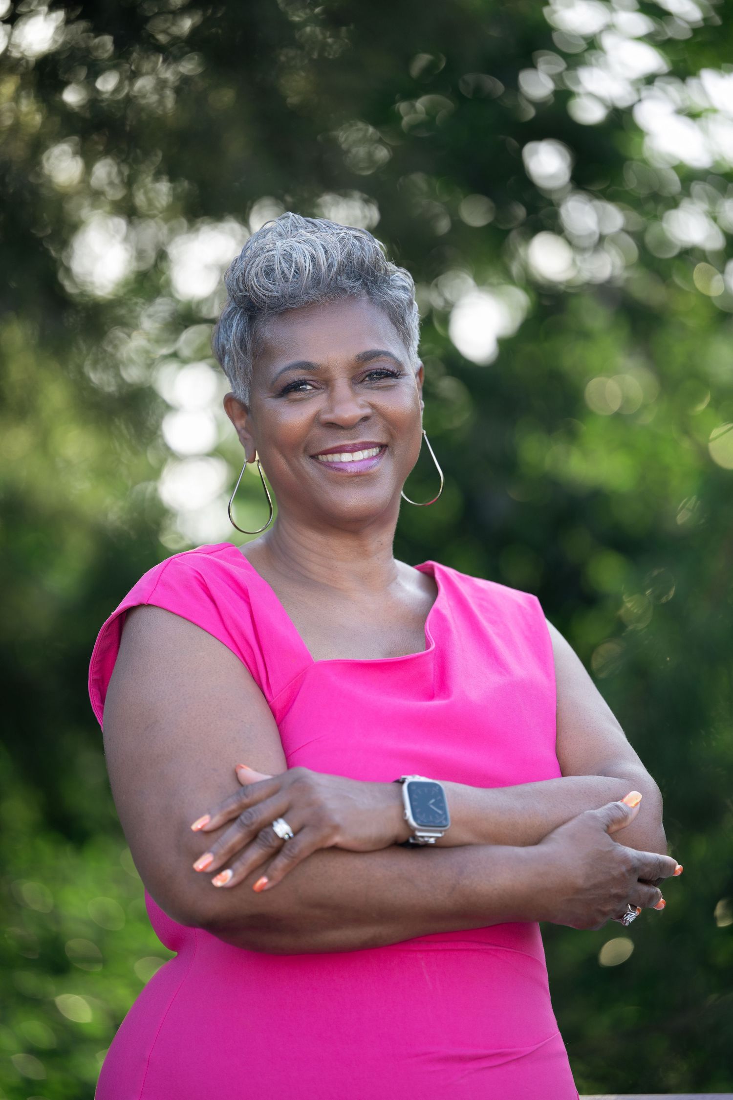 Confident smiling Black woman with gray hair wearing pink dress posing outdoors with arms crossed.