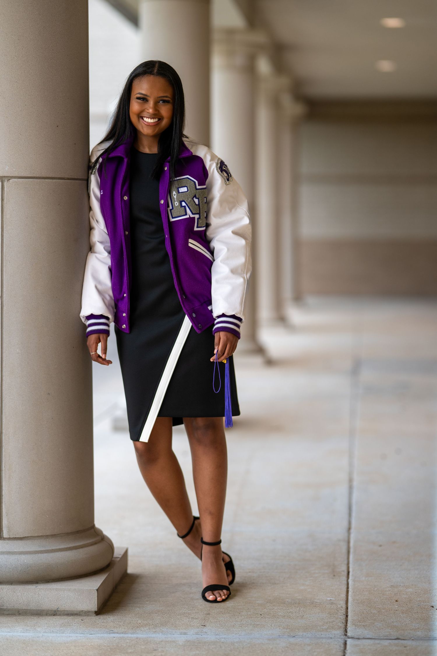 Smiling young woman in a purple and white varsity jacket and black dress, leaning against a column.