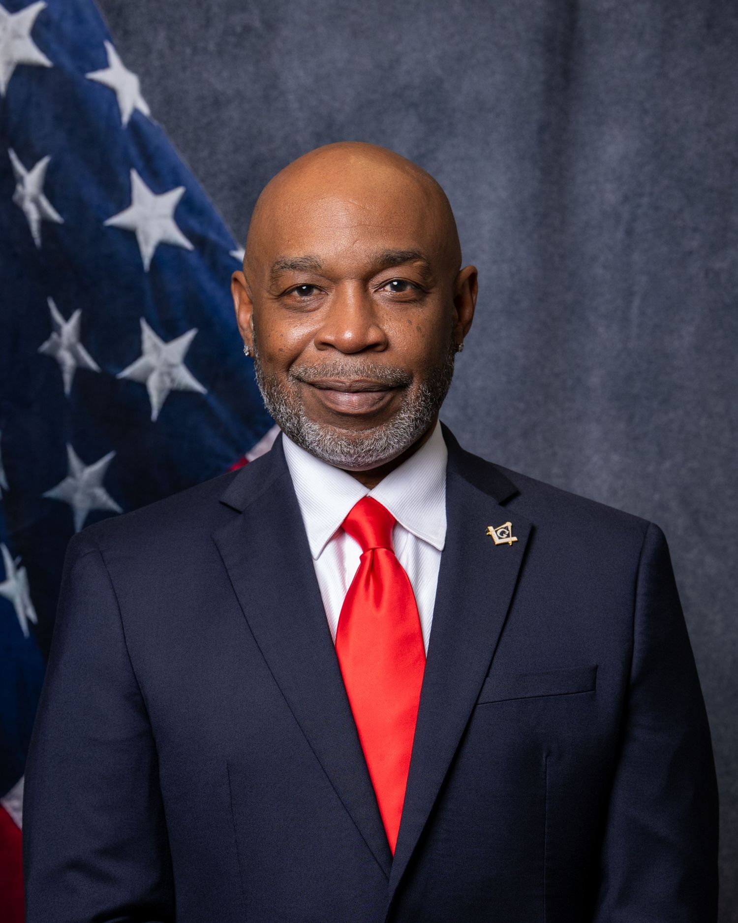 Professional headshot of a man in a navy suit and red tie posed before an American flag.