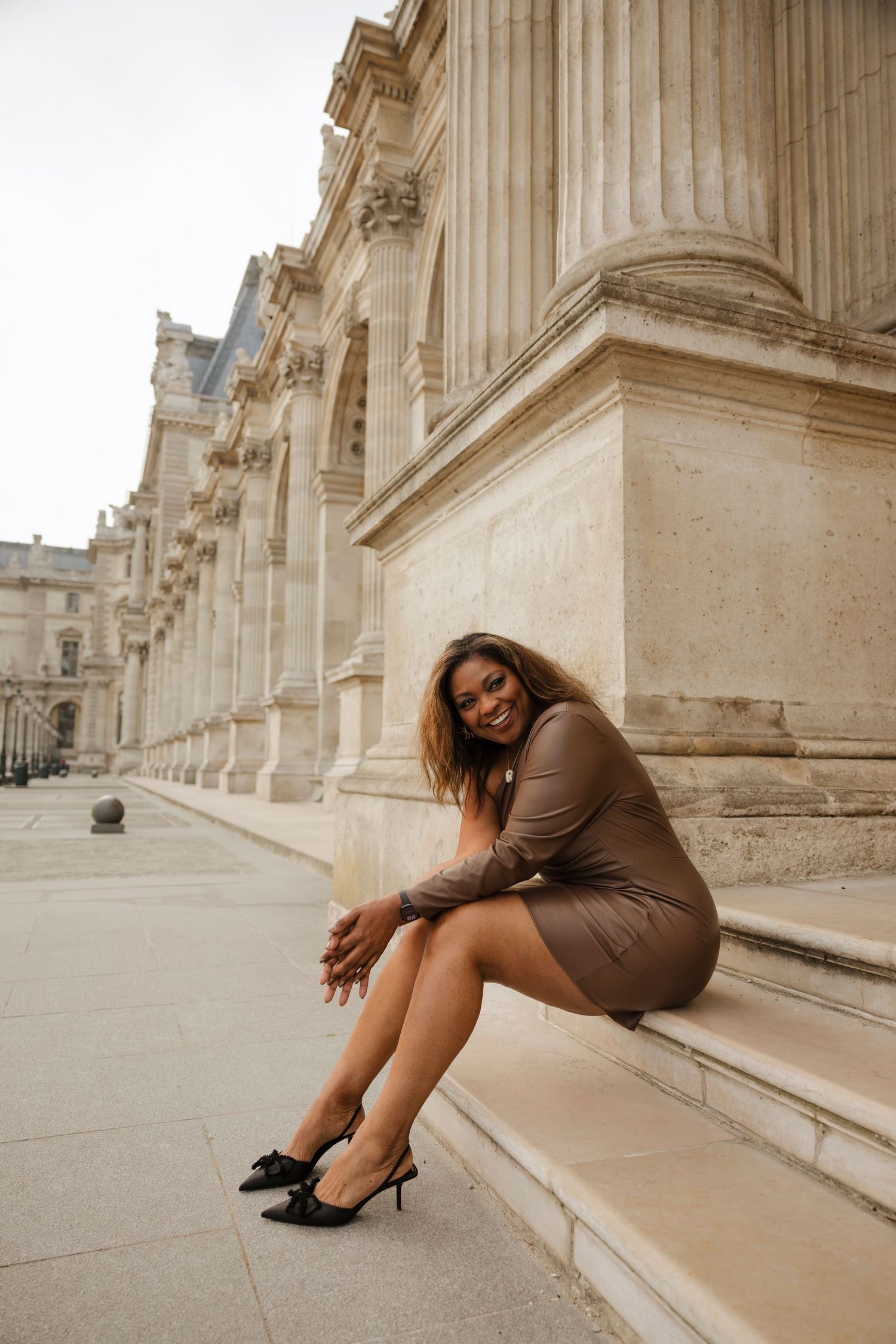 A smiling woman in a brown mini dress and black heels poses on steps outside the Louvre Museum in Paris.