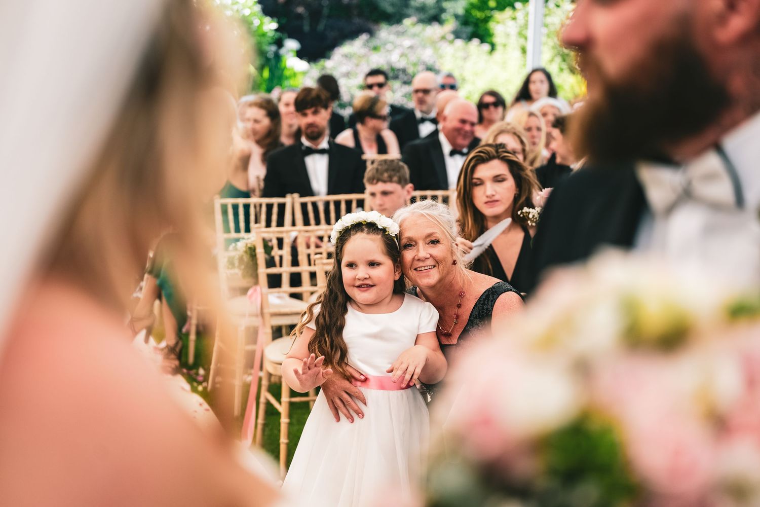 A smiling flower girl in white dress stands with grandmother during outdoor wedding ceremony.
