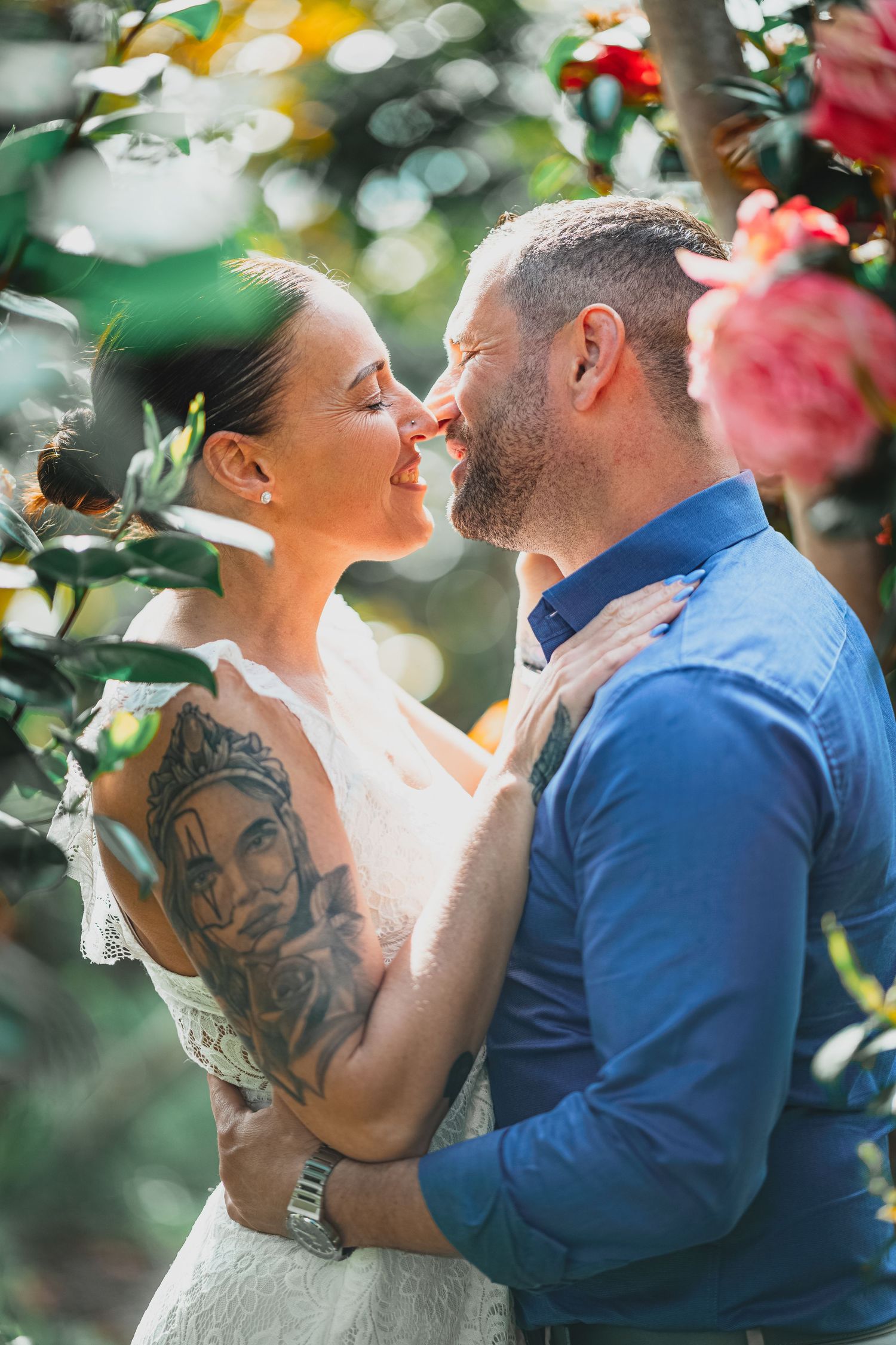 Tattooed bride and groom share a tender nose-to-nose moment surrounded by blooming roses.