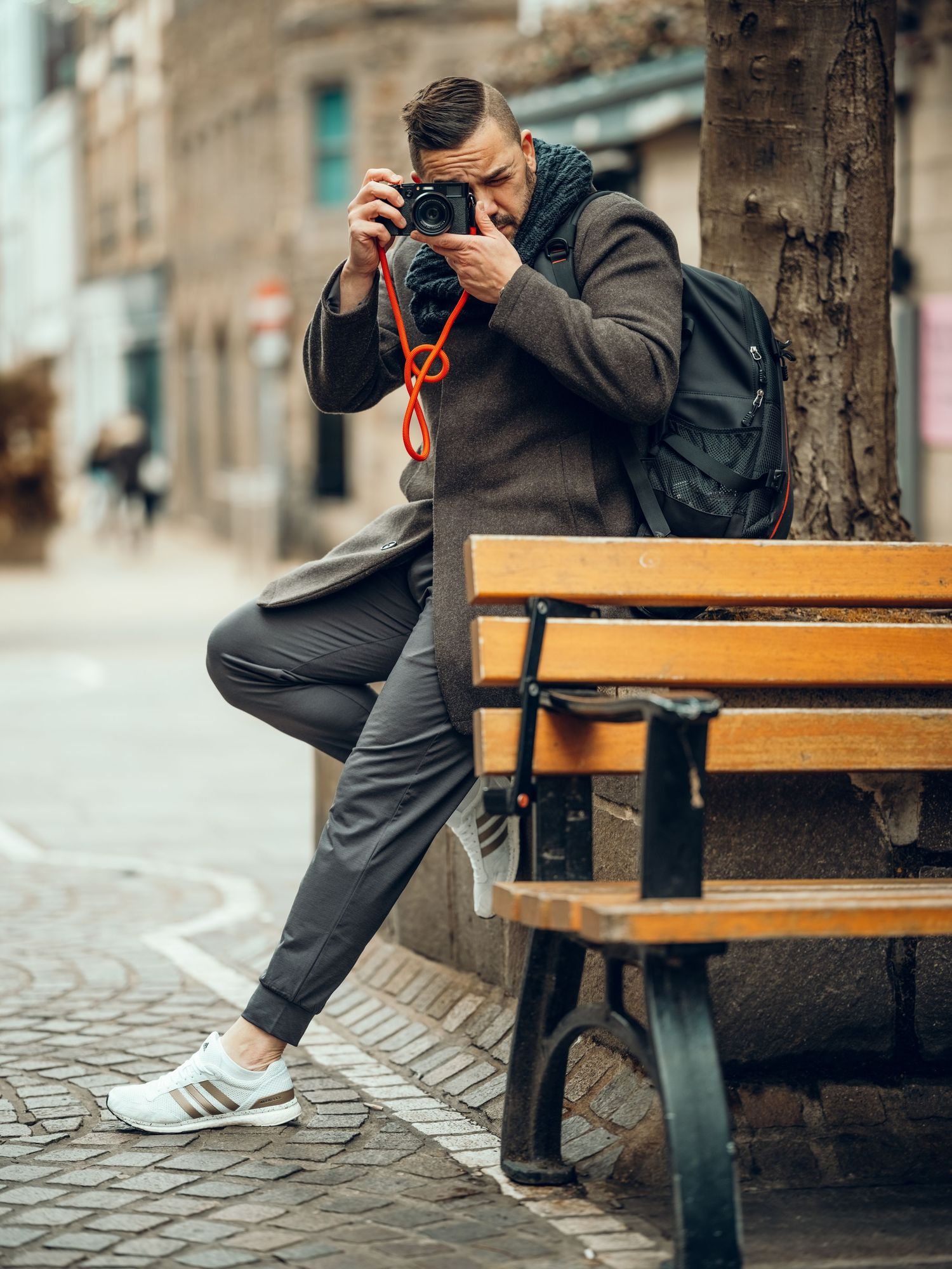 A stylish man photographs with a camera on a cobblestone street, wearing a brown coat and white sneakers near a wooden bench.