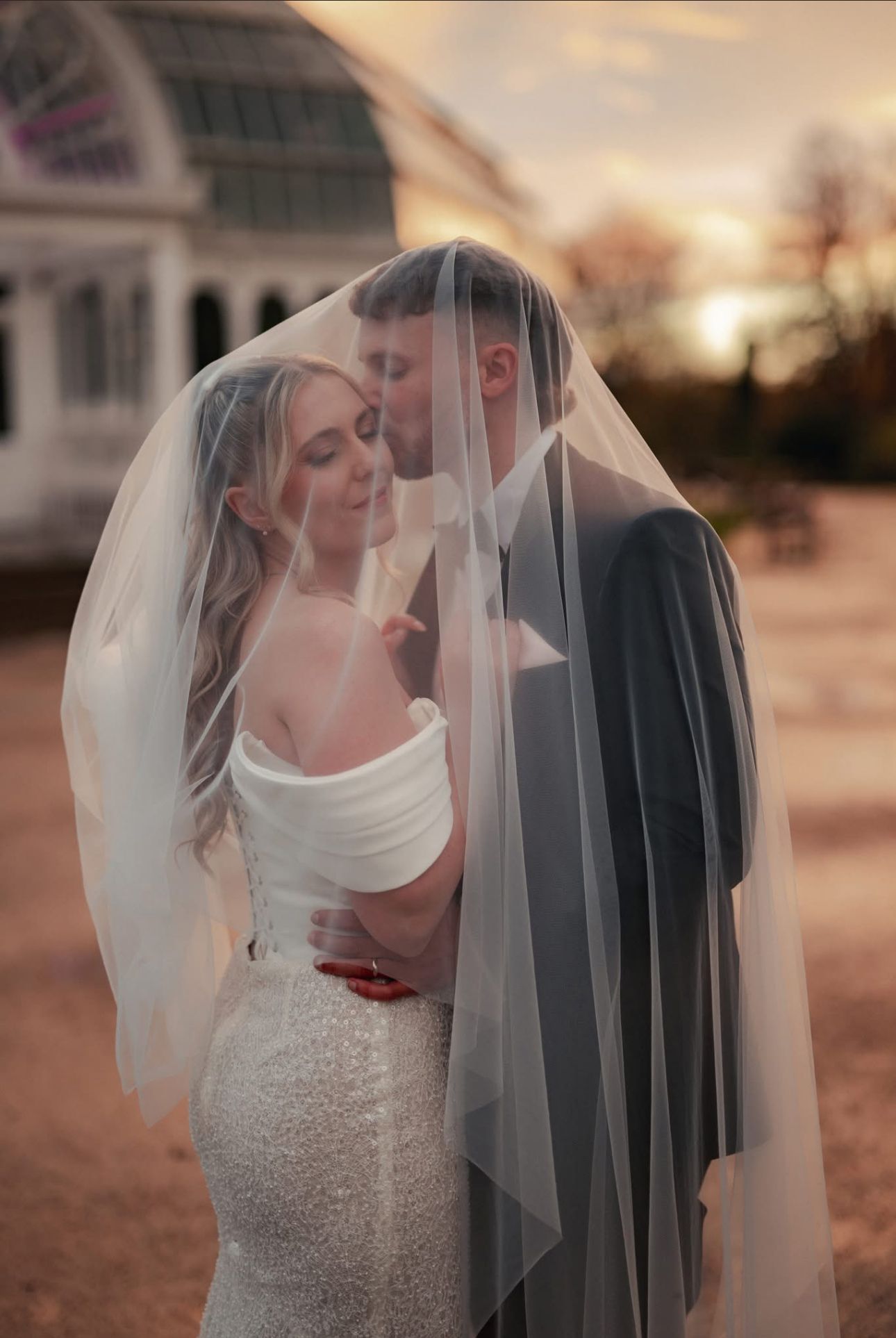 Bride and groom share intimate moment under sheer veil at sunset with greenhouse backdrop.