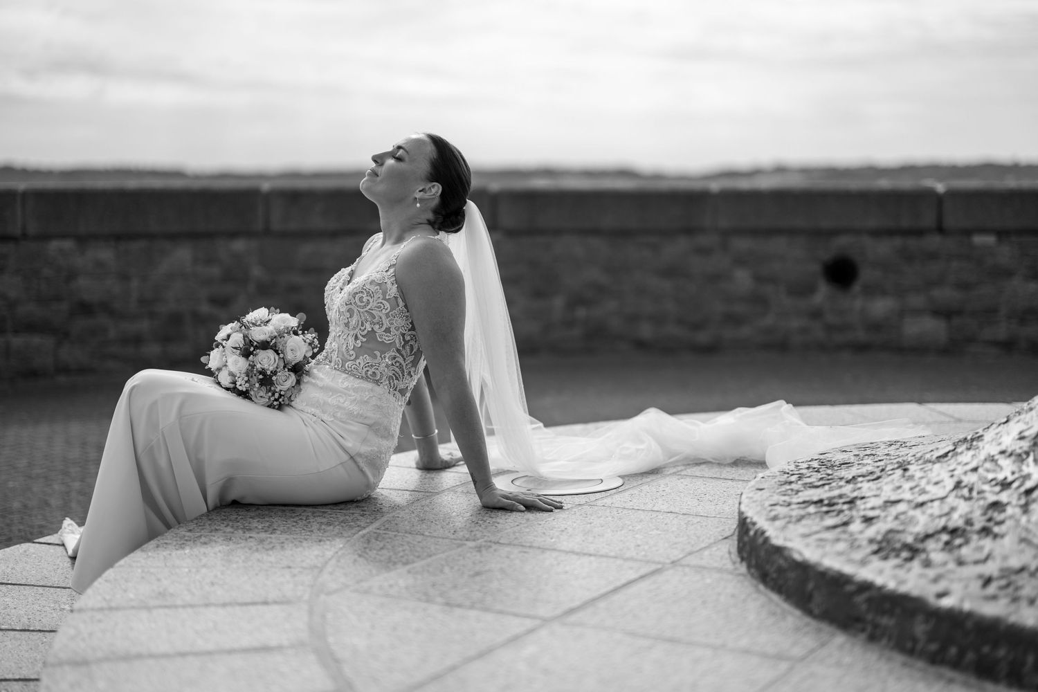 Bride in white gown sits gracefully on stone pavement holding bouquet with veil flowing behind her.