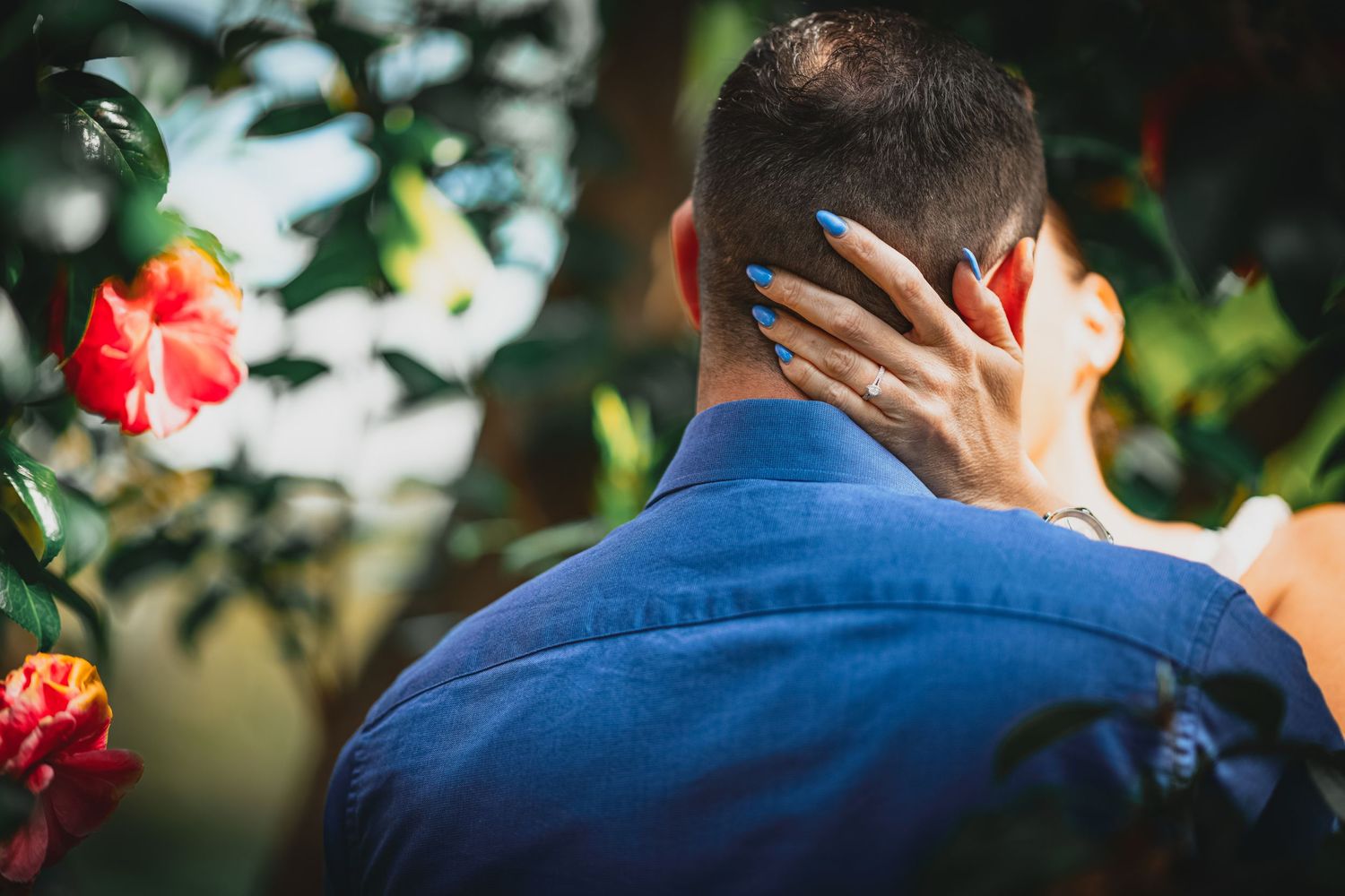Close-up of woman's hand with blue nails gently touching man's face amid vibrant red tropical flowers.