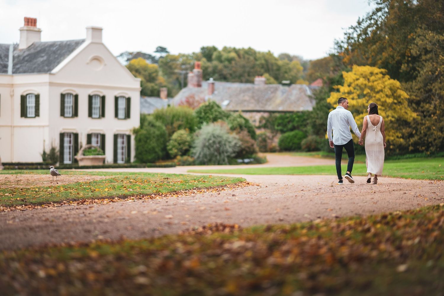 Couple walking hand in hand along a gravel path toward an elegant white country estate in autumn.