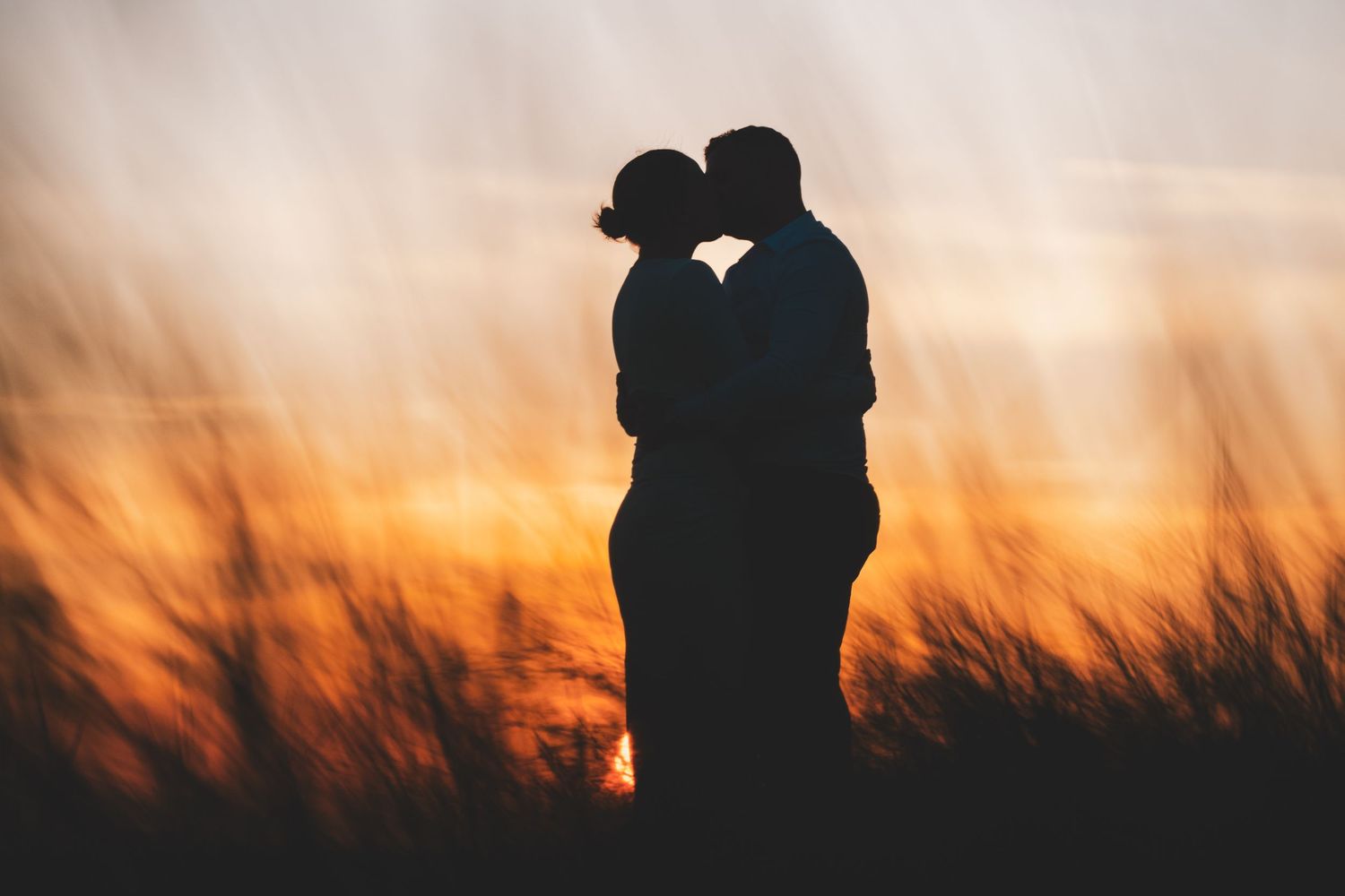 Silhouette of couple sharing a kiss at sunset with dramatic orange sky and tall grass backdrop.