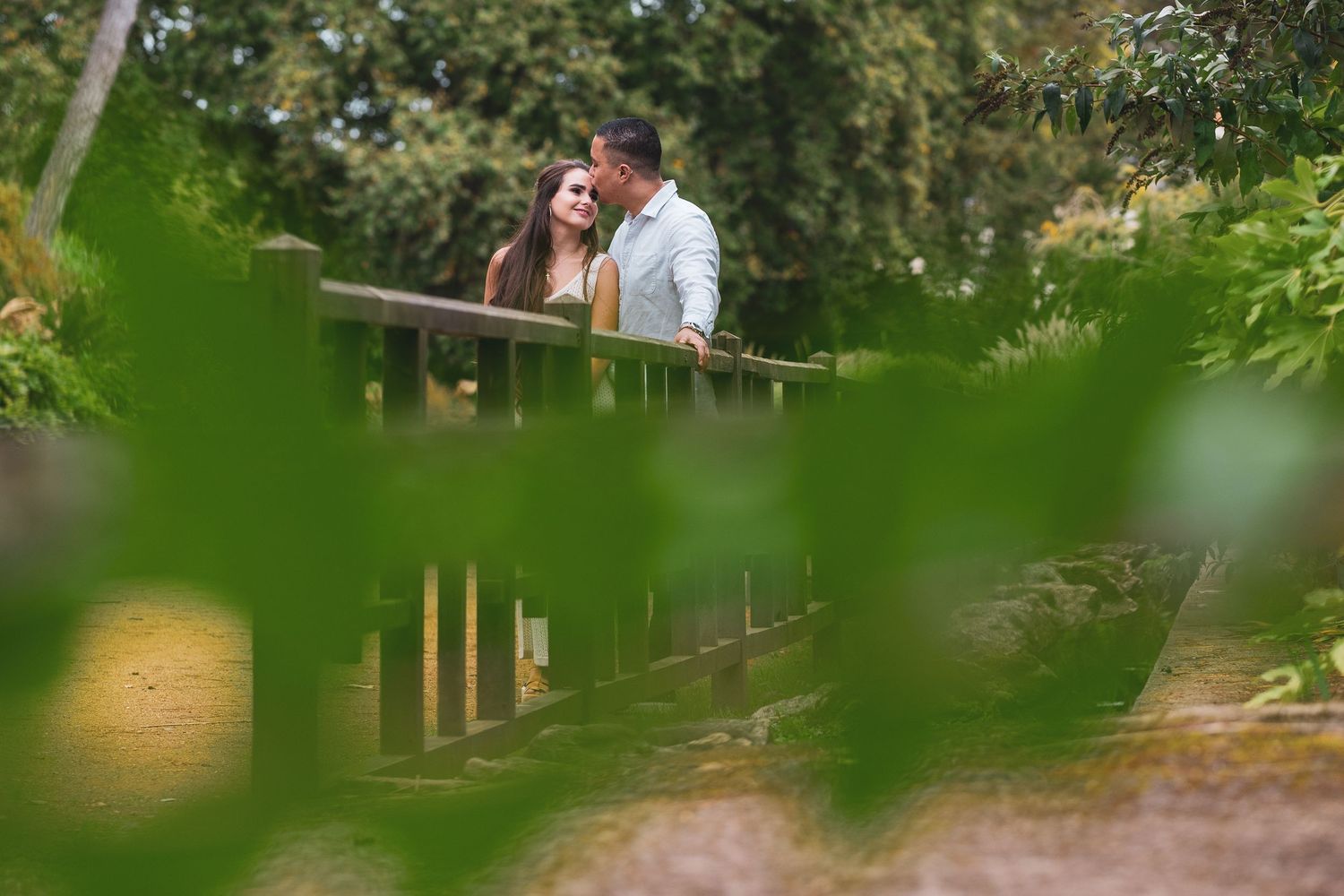 Couple sharing a tender moment on a wooden bridge surrounded by lush green garden foliage.