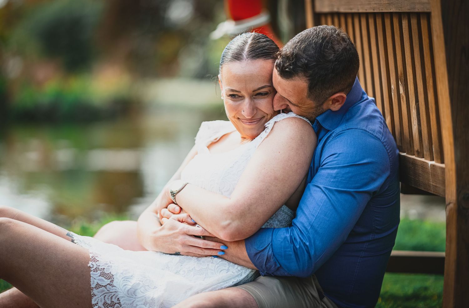 Couple embracing tenderly on a bench beside a tranquil pond during an outdoor engagement photo session.