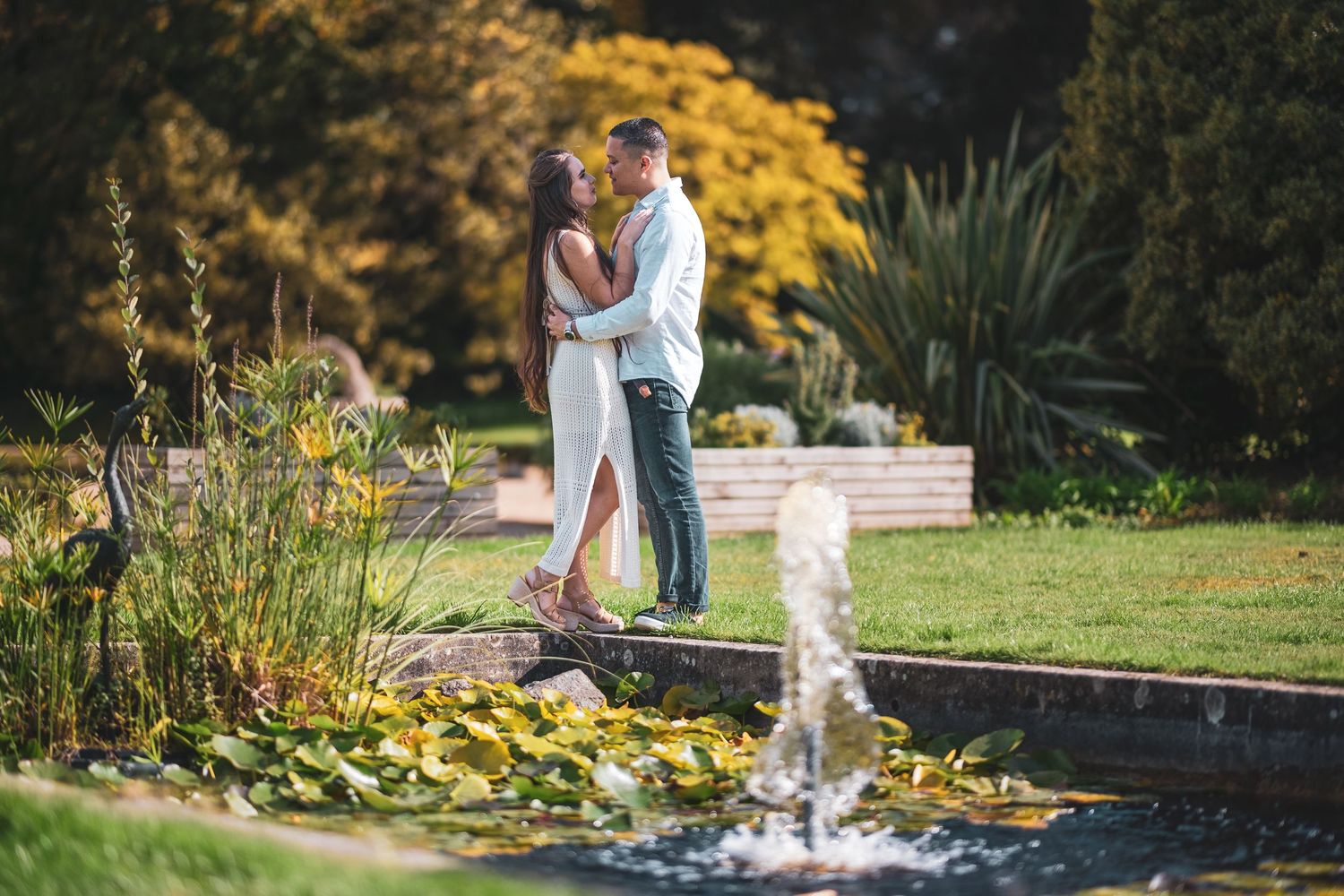 Couple embracing romantically beside a garden fountain surrounded by lush autumn foliage and greenery.