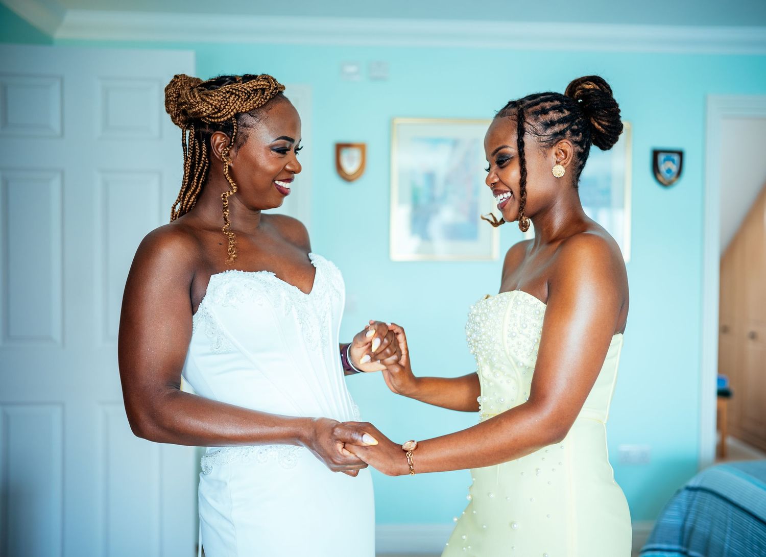Two women in white dresses smile and hold hands in a bright blue room during wedding preparations.